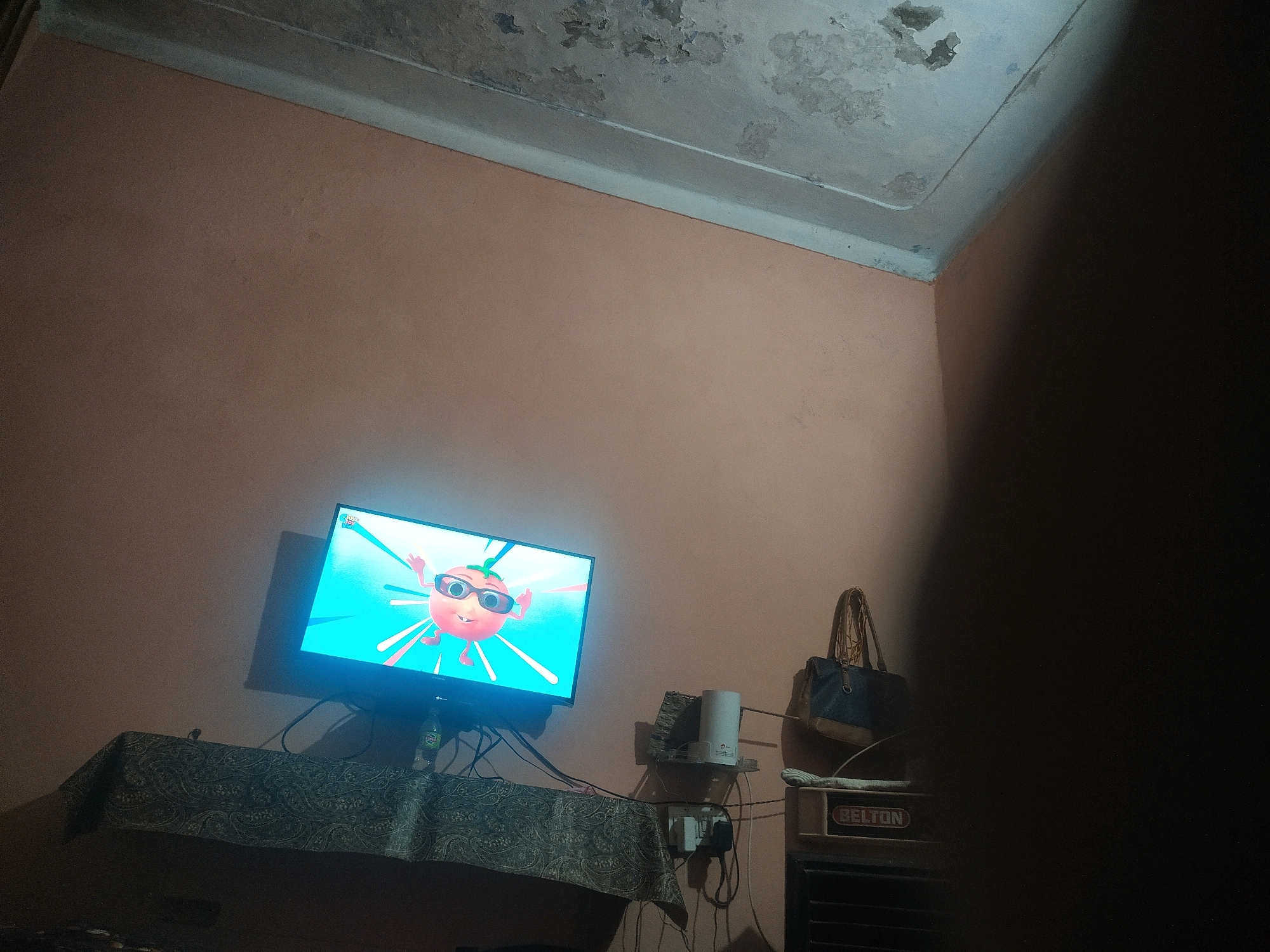 The image depicts an indoor scene, likely a living room, in Saharanpur, India. A flat-screen television is mounted on the wall, displaying a cartoon character wearing sunglasses against a bright blue background with radiating lines. The TV is placed on a table covered with a dark patterned cloth. To the right of the TV, a white cylindrical object, possibly a router or humidifier, sits on a small shelf attached to the wall. Below it, a power strip with multiple plugs is visible. Further to the right, a handbag with brown straps and a blue body hangs on the wall. Adjacent to the handbag, a metallic grille with the word "BELTON" in red lettering is present, possibly part of an appliance like an air cooler. The walls are painted a pale peach color, and the ceiling shows signs of wear and discoloration. The overall lighting is dim, with a shadow obscuring the right side of the image.