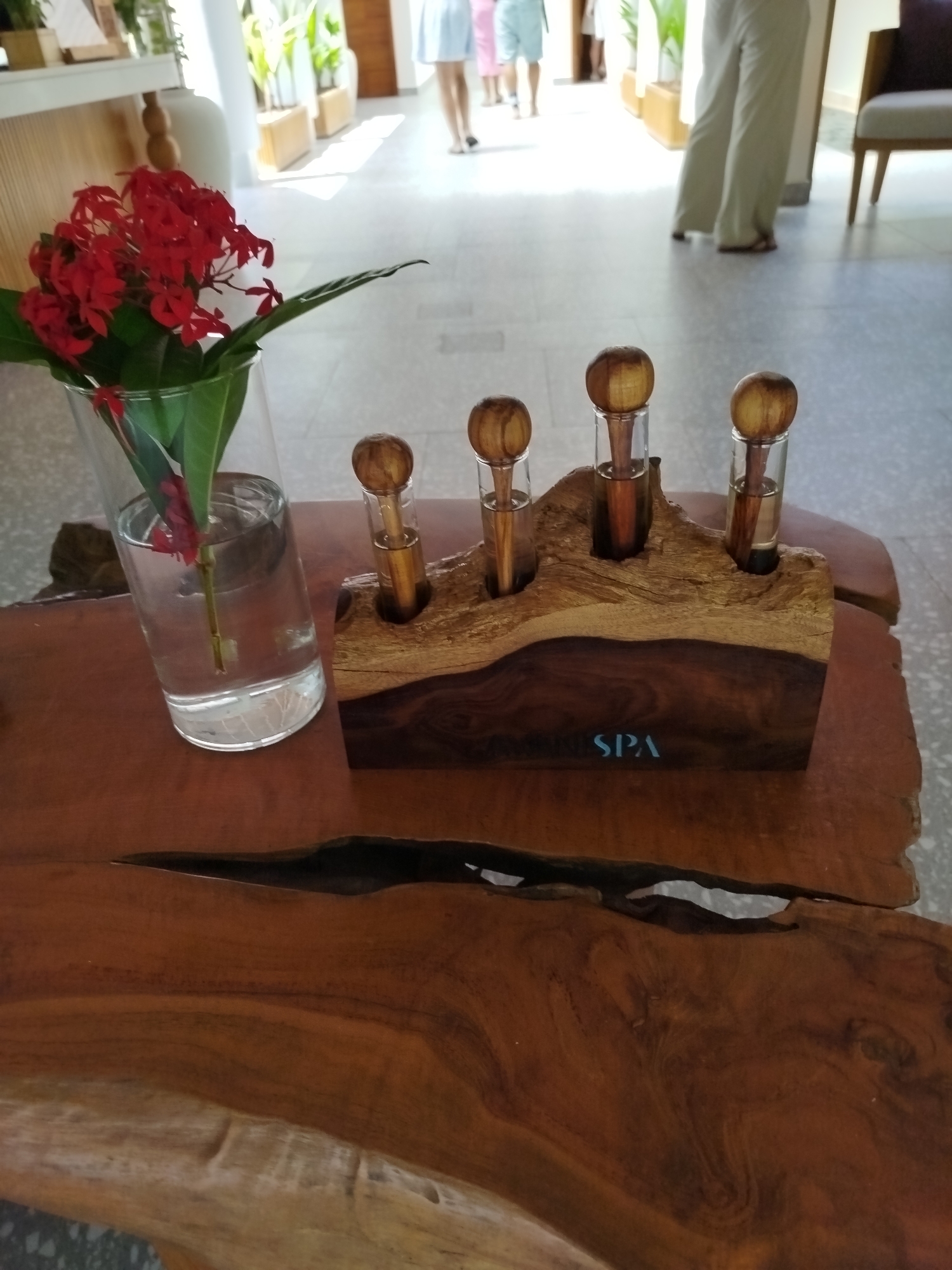The image depicts a close-up view of a wooden table in what appears to be a reception or common area of a resort in Kendhoo, Maldives. On the table, a clear glass vase holds a bunch of vibrant red flowers, possibly Ixora, with green leaves. To the right of the vase is a rustic, carved wooden stand with four small glass vials, each containing a clear liquid and capped with a wooden sphere. The word "SPA" is subtly etched in light blue on the front of the wooden stand. In the background, the scene is softly lit by natural light, with glimpses of other resort elements like furniture, potted plants, and the blurry figures of people walking. The overall impression is one of natural beauty and relaxation, characteristic of a tropical island destination.