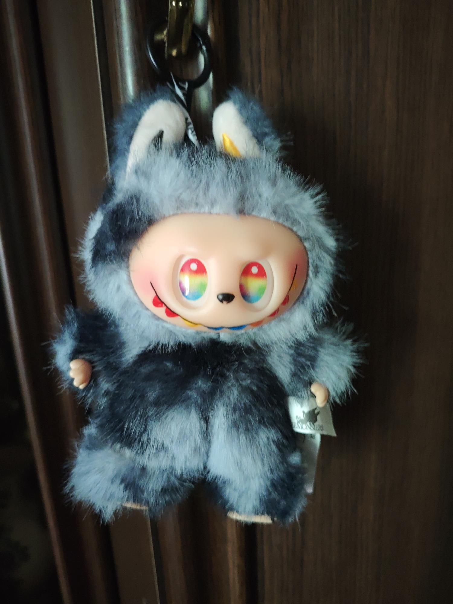 The image shows a plush toy with a furry gray and black suit, large head, and round, rainbow-colored eyes. The toy is hanging from a hook against a dark brown wooden door. The toy has small, beige hands and feet, and a wide smile with red lips and visible teeth. A small white tag is attached to the toy's right arm. The background consists of the textured surface of the door and a hint of a curtain on the left. The scene is indoors. There is no indication of the city of Stavropol, Russia.