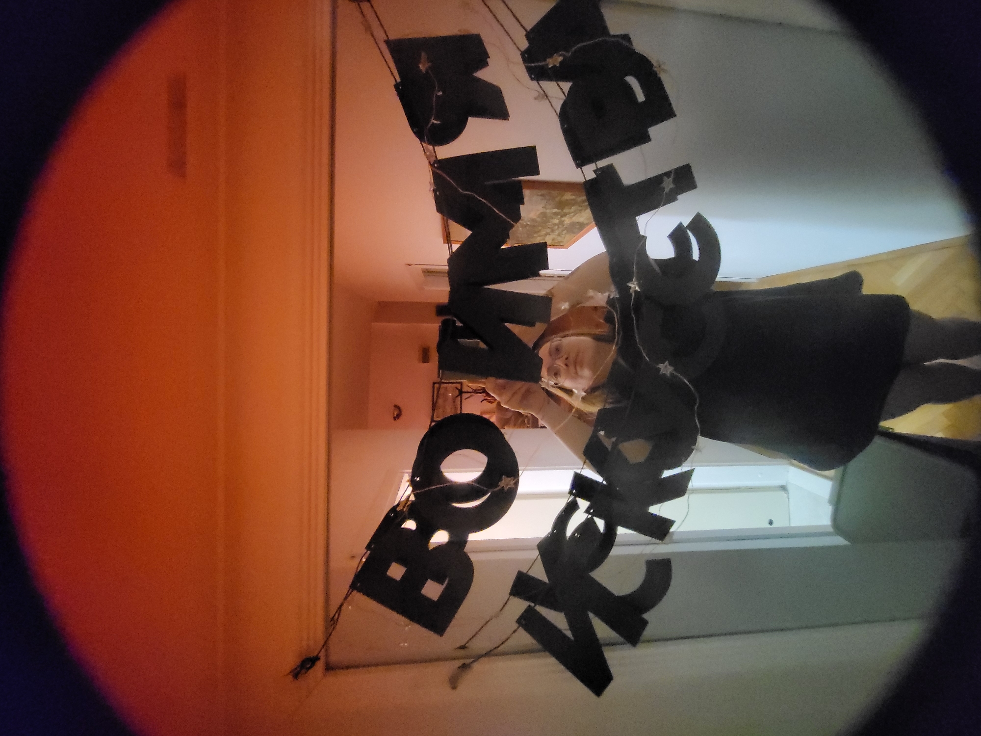 A person wearing a witch costume stands in a room decorated with a "Boo to You" banner. The room has orange and white walls. The location is not discernible from the image.