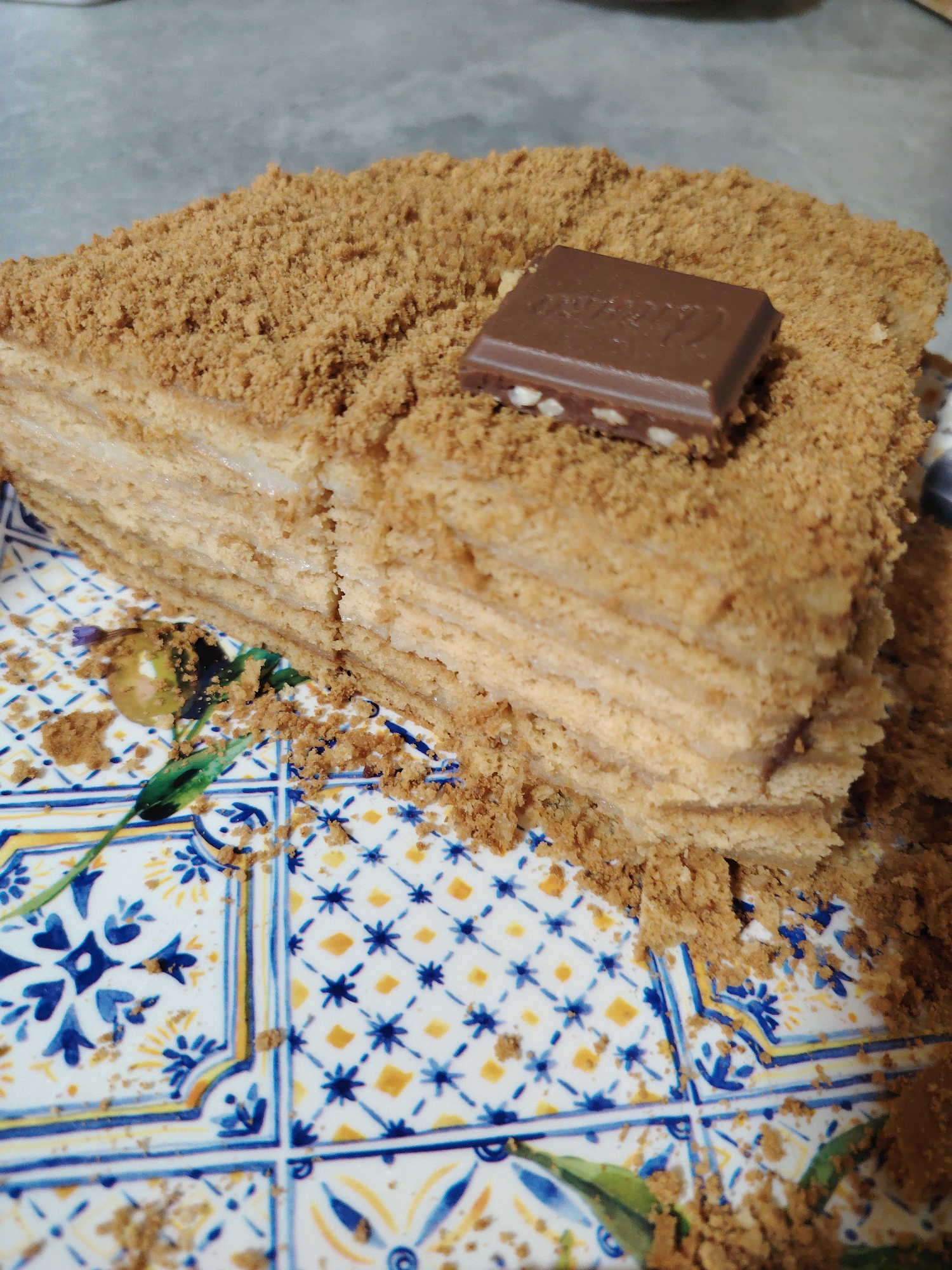 A close-up, eye-level shot shows a slice of honey cake on a decorative plate. The cake is layered with thin, light brown cake wafers and a pale cream filling, and it is generously coated with crushed cake crumbs on top and sides. A small, rectangular piece of milk chocolate with visible nuts sits on the top crumb layer, slightly askew. The plate has a vibrant blue and white patterned design with yellow accents, featuring floral and geometric motifs. Crumbs have fallen from the cake onto the plate. The background appears to be a grey, textured surface, possibly a table. The scene is brightly lit, emphasizing the textures and details of the cake and plate. There is no indication of a specific location in Rostov-on-Don, Russia.