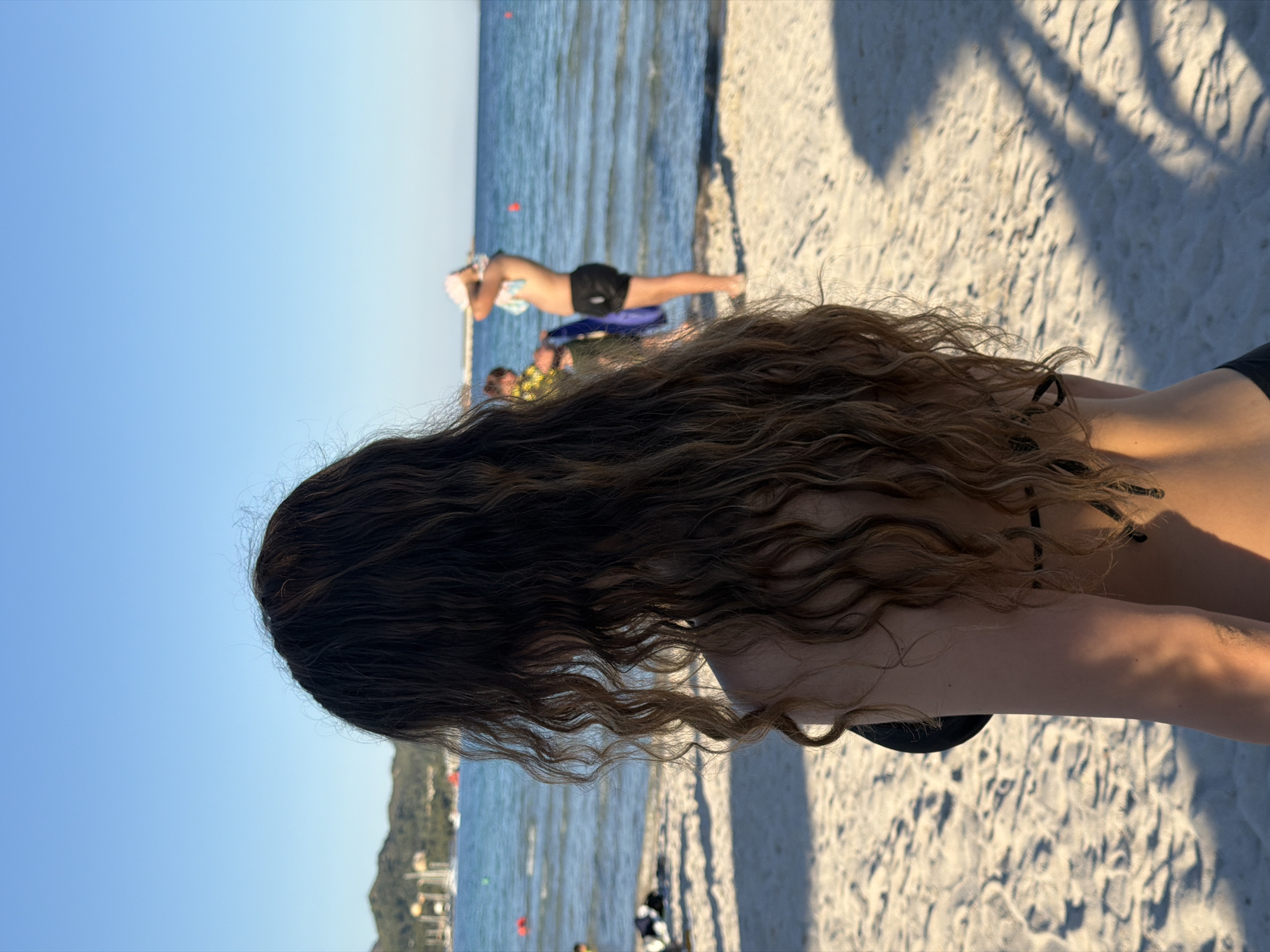 A sunlit beach scene in Alcúdia, Spain. The back of a person with long, wavy brown hair dominates the foreground, wearing a black bikini top. The scene stretches from the person towards the horizon with a man lying on a blue surfboard, other people visible in the water. The shoreline and distant buildings are present. The beach's sand displays shadows from trees or foliage. The sky is a clear blue, while the water color varies from light to dark blue.