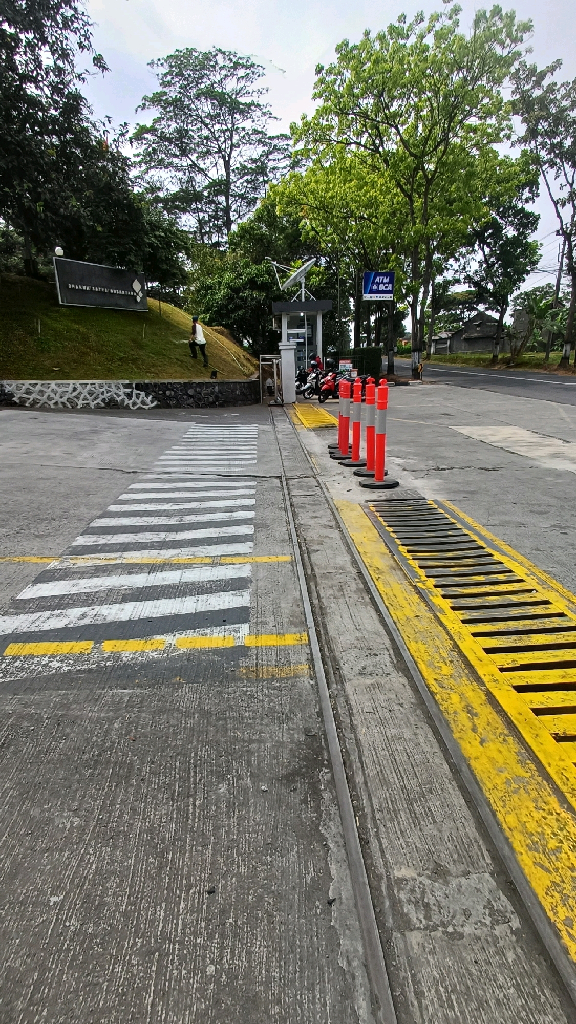 The image captures a view of a paved area with a zebra crossing painted on the ground. Yellow lines are painted along the edge of the zebra crossing, and a row of orange and white bollards stands to the right of the crossing. Beyond the bollards, there is a metal grate. A small security booth with an ATM sign is visible in the background, with motorcycles parked nearby. A sign with text that appears to read "DHARMA SATYA NUSANTARA" is situated on a grassy incline to the left. The scene is framed by trees and an overcast sky, suggesting an outdoor environment. The image is taken at the entrance to the Dharma Satya Nusantara complex in Kranggan, Indonesia.