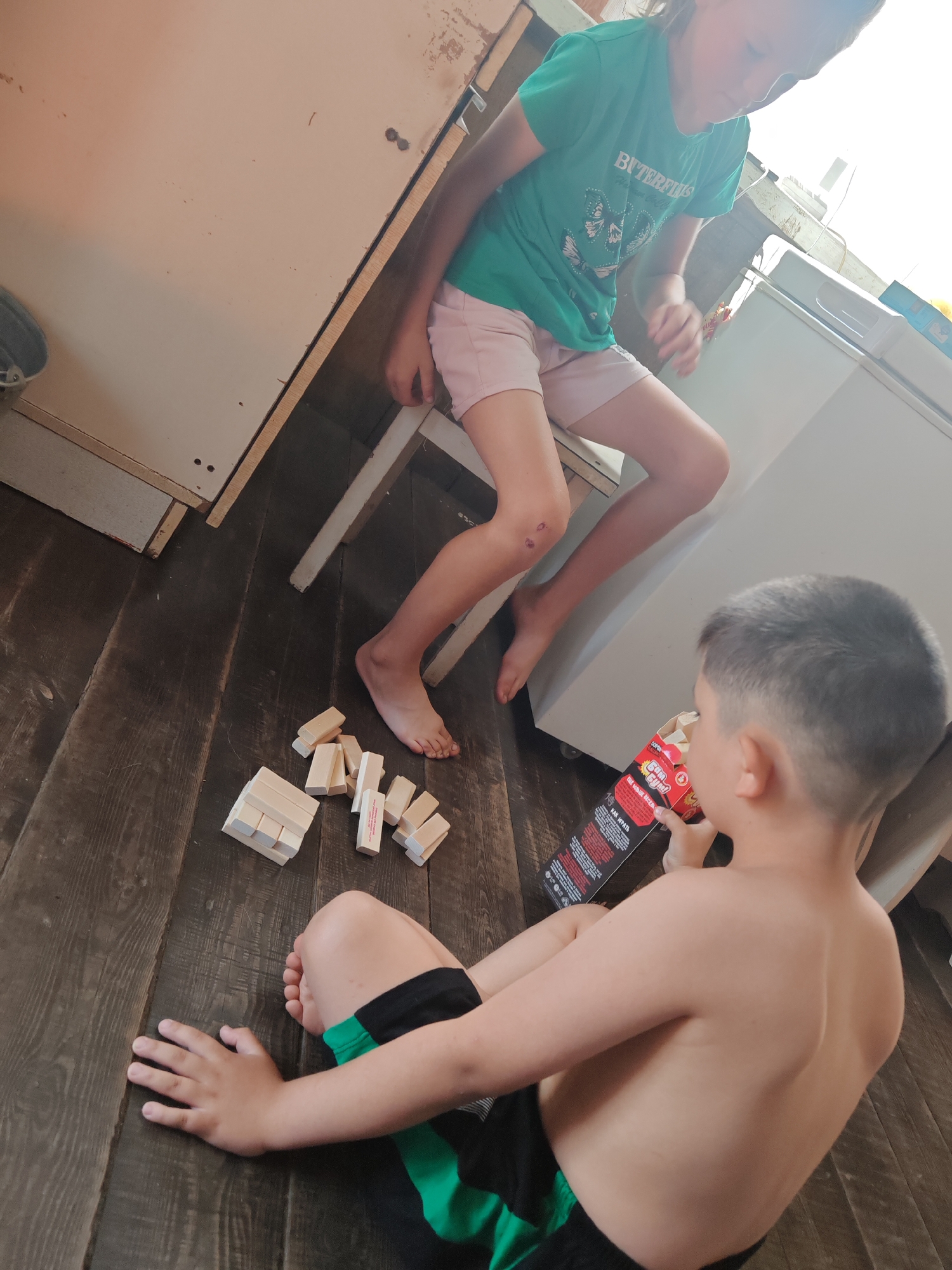 The image shows two young children playing a game with wooden blocks on a wooden floor, likely in a home in Chernakovo, Russia. On the left, a girl wearing a green t-shirt and pink shorts is sitting on a stool. Her t-shirt has an image of butterflies and text that reads "BUTTERFLIES". She has a scraped knee. On the right, a boy is sitting on the floor, facing away from the camera, shirtless and wearing black and green shorts. He is holding a box with red and black lettering, which appears to be a game or snack. Scattered on the floor around the children are several wooden blocks, some stacked in a small tower, suggesting they are playing Jenga or a similar block-stacking game. The lighting suggests it is daytime. The overall scene is casual and domestic.