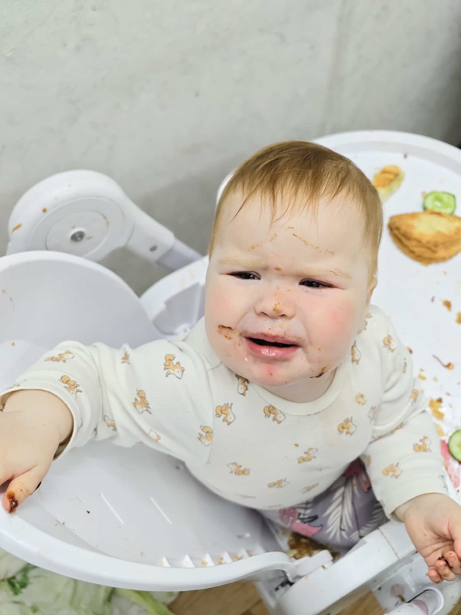 A baby is sitting in a white highchair. The baby has fair skin, light brown hair, and a face covered in food. The baby appears to be crying, with red eyes and a downturned mouth. The baby is wearing a long-sleeved white shirt patterned with small fox figures, and patterned pants are visible underneath. The highchair tray contains remnants of food, including what appears to be bread and cucumber slices. The background is a plain off-white wall. Based on context, the location is Chaikovsky, Russia.