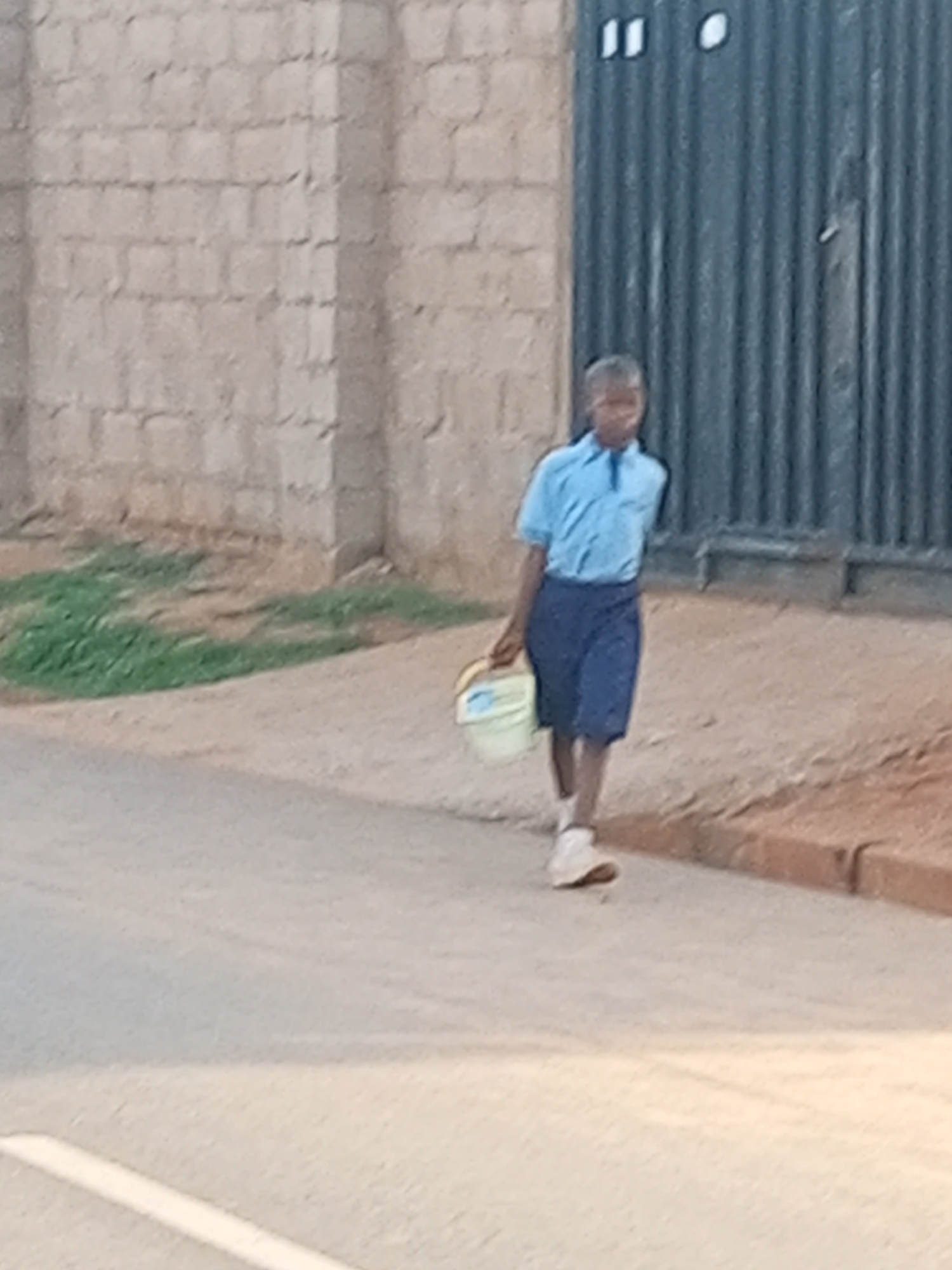 A young boy walks on a paved road in Tempure, Nigeria, carrying a yellow bucket in his right hand. He is wearing a light blue short-sleeved shirt and dark blue shorts, with white socks and light-colored shoes. The road is bordered by a dirt path and then a wall made of beige bricks on the left, and a corrugated metal gate on the right. Green grass is visible on the embankment between the road and the brick wall.