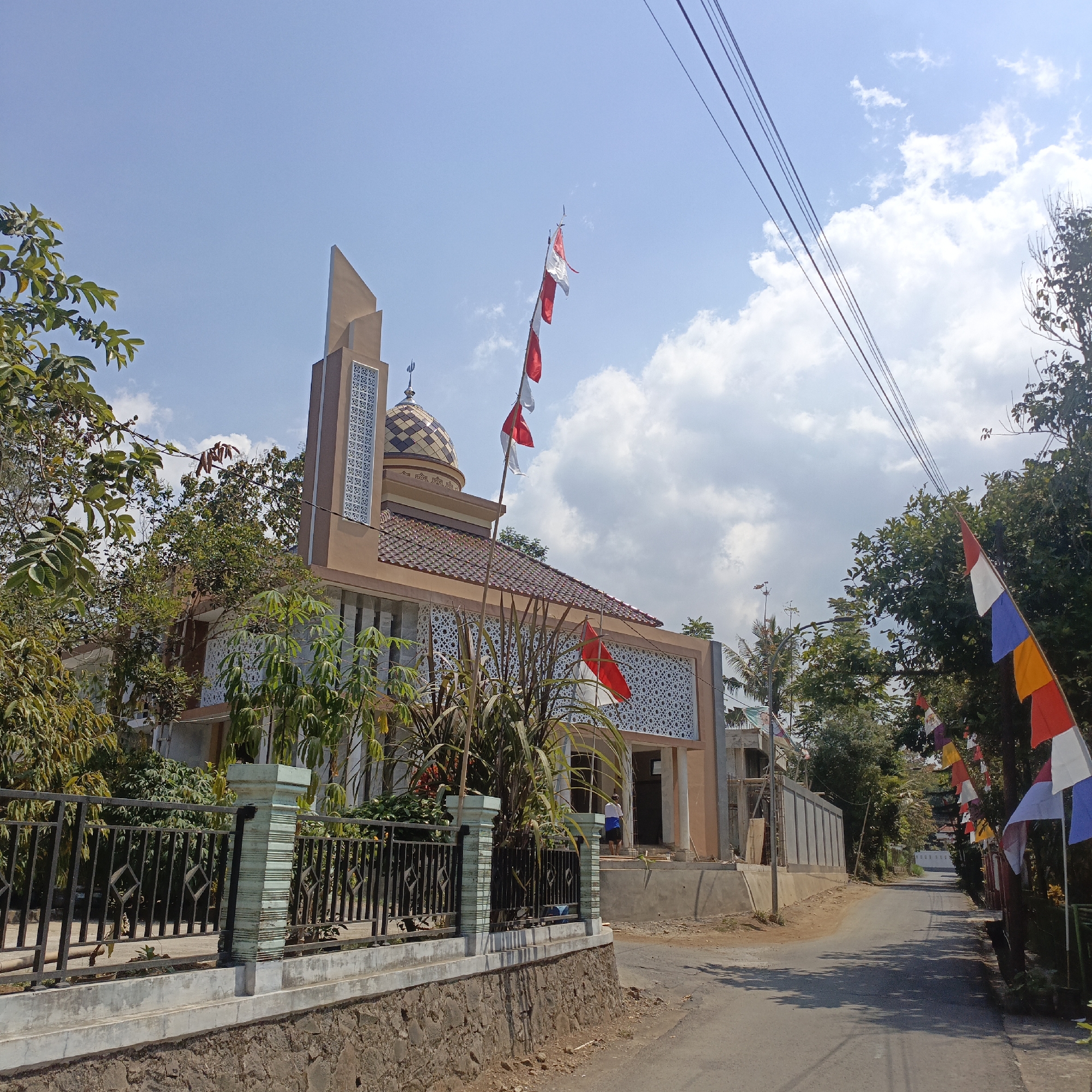This is a street-level, eye-level, medium shot of a mosque with a tan facade and a domed roof, located in Salatiga, Indonesia. The mosque features a tall, slender minaret with decorative patterns. Several Indonesian flags, red and white, are displayed on poles, and a string of colorful flags decorates the street on the right side of the image. Trees and foliage surround the building, and overhead power lines are visible against a bright blue sky with scattered clouds. The street is paved and appears to be on a slight incline, with a low stone wall and a black metal fence in the foreground. One person in a blue and white shirt is standing near the entrance of the mosque, possibly a construction worker, as scaffolding is visible on the side of the building. The overall scene suggests a bright, sunny day, likely during a festive period given the flags.