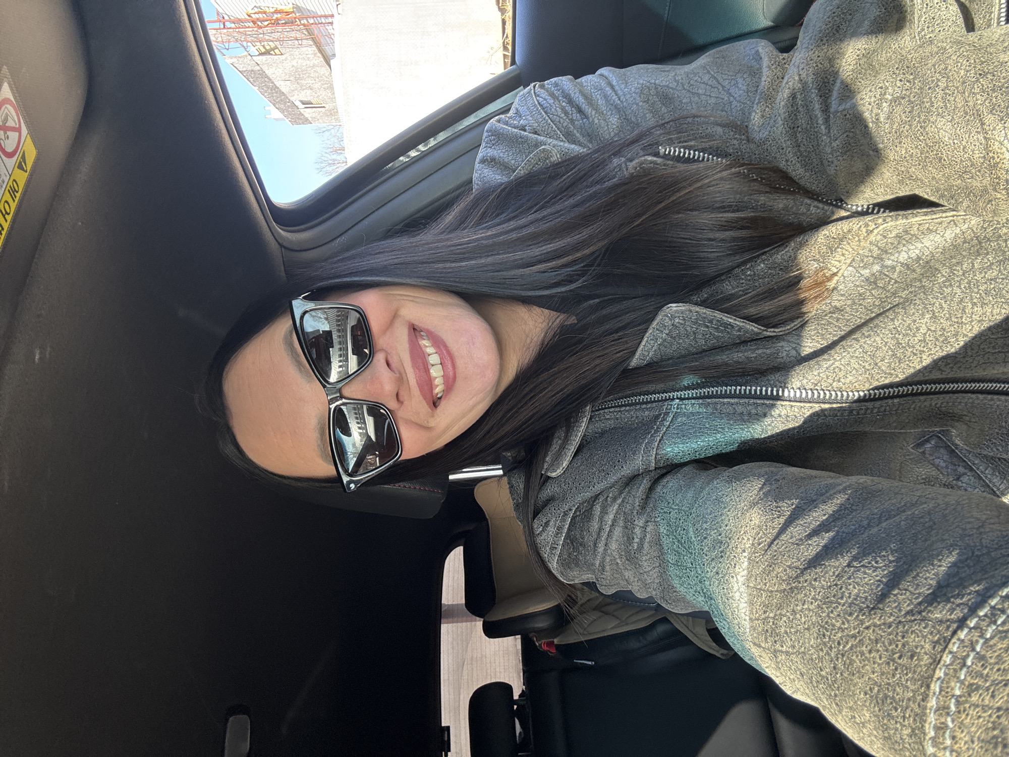 A woman with long dark hair and wearing sunglasses and a gray leather jacket is sitting in a car. The car appears to have an open sunroof. In the background, through the sunroof, a building under construction with scaffolding is visible. The location is identified as the city of Chaikovsky, Russia.