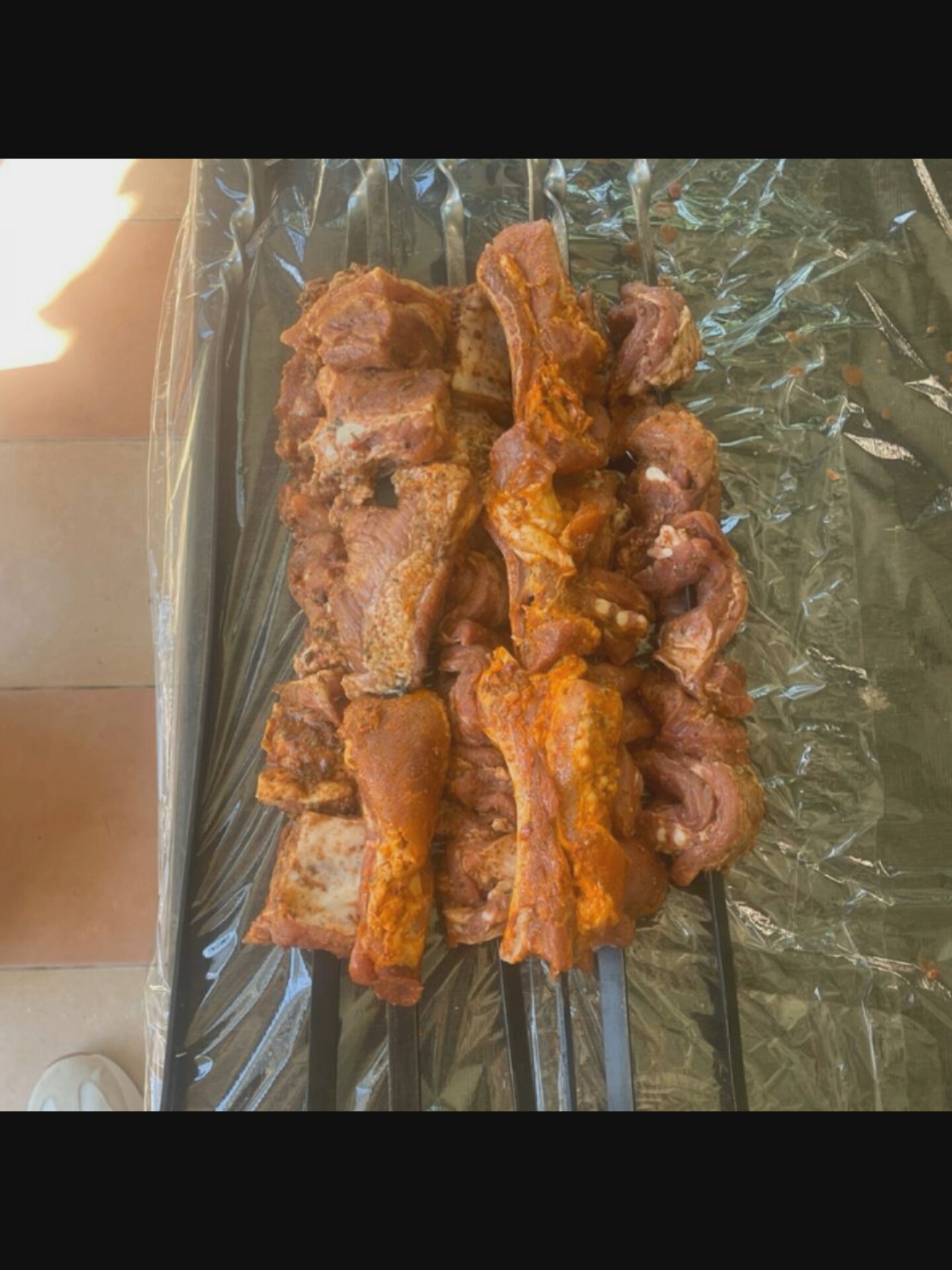 The image shows a close-up, overhead view of raw meat skewers. The meat appears to be marinated in a reddish-orange spice blend and is arranged on metal skewers. The skewers are placed on a dark, crinkled surface, possibly a disposable tablecloth or plastic sheeting, which itself rests on a black, slatted surface, likely a grill or a warming rack. In the background, to the left, there is a hint of an outdoor setting with a tiled surface in shades of brown and an indication of sunlight creating a shadow. The lighting suggests it is daytime. No people are visible in the frame, and there is no visible text. The scene is entirely focused on the preparation of the meat. The overall impression is one of food preparation, likely for a barbecue or a similar cooking event. The location context is Bucharest, Romania.