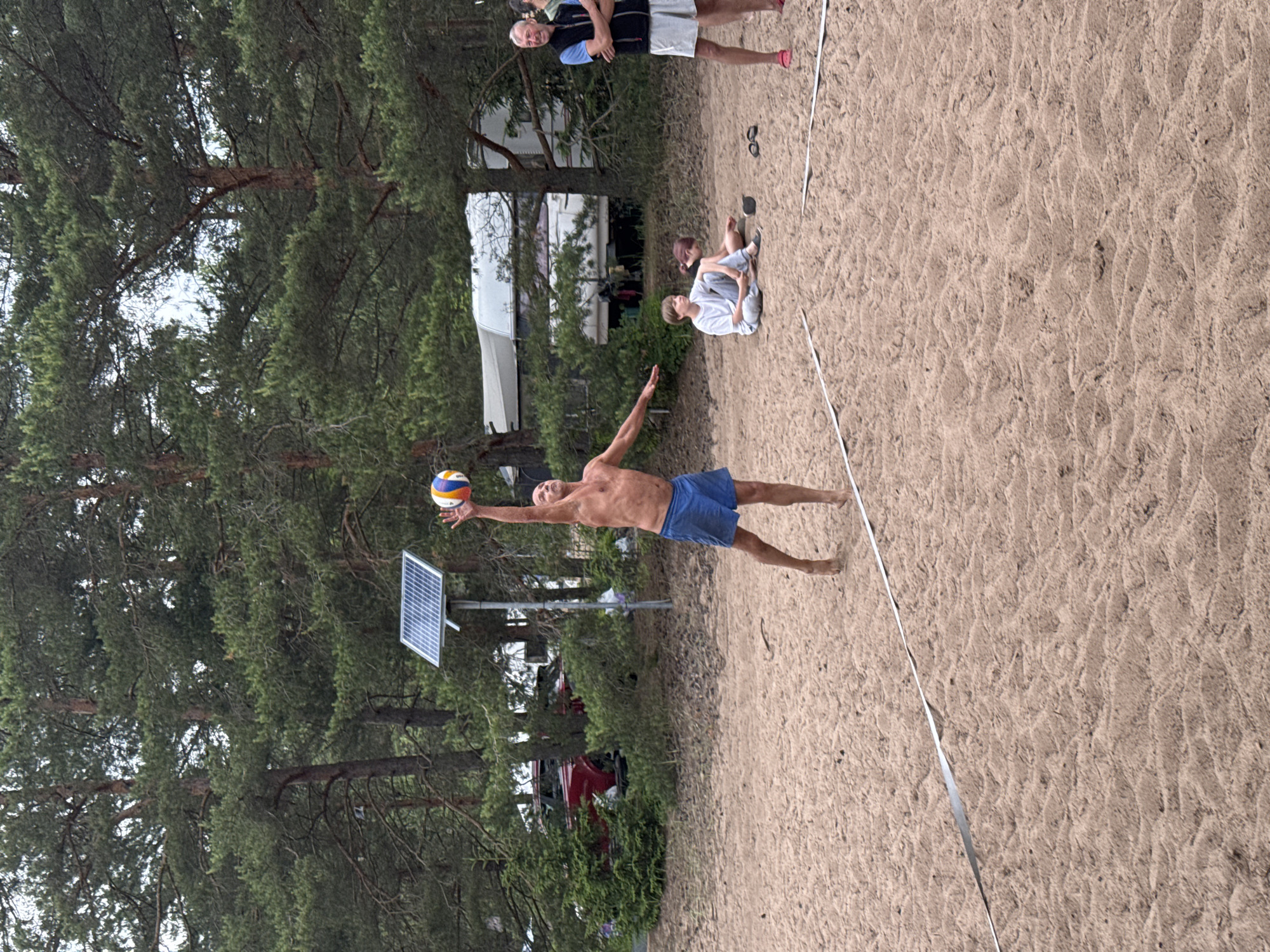 An outdoor, eye-level, full shot captures a group of people on a sandy beach, presumably in Zelenaya Roshcha, Russia, as indicated by the context. The foreground features a volleyball net marking a boundary on the sand. A man, bare-chested and wearing blue shorts, is in the process of hitting a volleyball, which is suspended in the air above him. His body is angled, with one arm extended upwards towards the ball and the other outstretched to his side. His legs are bent, suggesting motion. In the midground, two children are seated on the sand. One, a young boy in a grey t-shirt and pants, sits with his legs extended. Beside him, a person with dark hair is also seated, facing away from the camera. Further back, two adults stand; one man in a black vest over a blue shirt and light shorts, arms crossed, and another man behind him. Behind the people, a line of trees, primarily pine, forms a dense backdrop. A red vehicle and a white RV are partially visible amongst the trees. A solar panel is mounted on a pole near the volleyball player. The overall lighting suggests an overcast day.