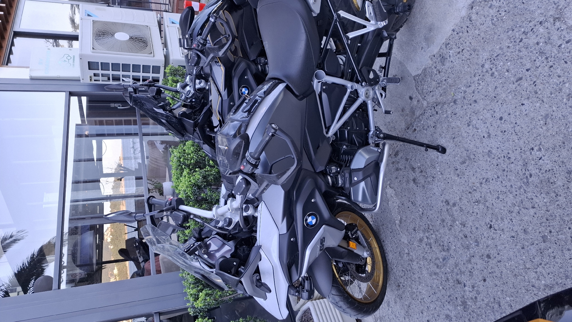 Two BMW R 1250 GS motorcycles are parked on a concrete surface in Boreti, Montenegro. The motorcycles are positioned next to a modern building with large windows reflecting a coastal view. Green shrubbery separates the motorcycles from the building. An air conditioning unit is visible on the building's exterior. The motorcycles are parked in an upright position with the side stand deployed.