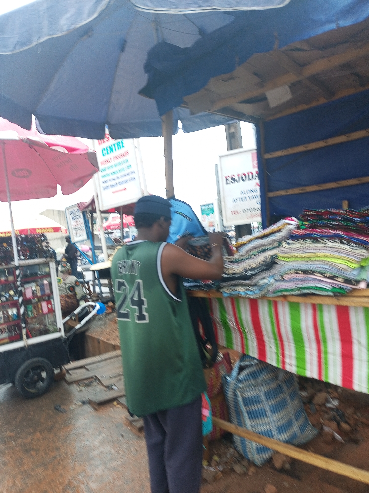 The image depicts an outdoor market scene, likely during the daytime, characterized by an array of vendor stalls operating under large umbrellas. In the foreground, a person is seen from behind, wearing a green sleeveless jersey with the number "24" on the back and dark trousers. This individual is standing at a market stall, seemingly sorting or arranging a large stack of colorful, folded fabrics or clothing items.
The stall itself has a wooden structure and is adorned with a vibrant striped fabric in red, white, and green patterns across its front. To the left, a mobile cart with wheels displays various small goods, such as watches and other accessories. A blue market umbrella provides overhead shade, and a pink umbrella is also visible further left. The ground is uneven and appears to be dirt or mud. Underneath the main stall, a large blue and white checkered woven bag rests on the ground.
In the background, several signs are visible. One white sign prominently displays "CENTRE" at the top, followed by "WEEKLY PROGRAM" and other text detailing activities or schedules, accompanied by an arrow. Another sign further back reads "ESJODA" at the top, followed by "ALONG", "AFTER YAW", and a telephone number starting with "TEL: 07065...". The scene conveys a bustling commercial environment.