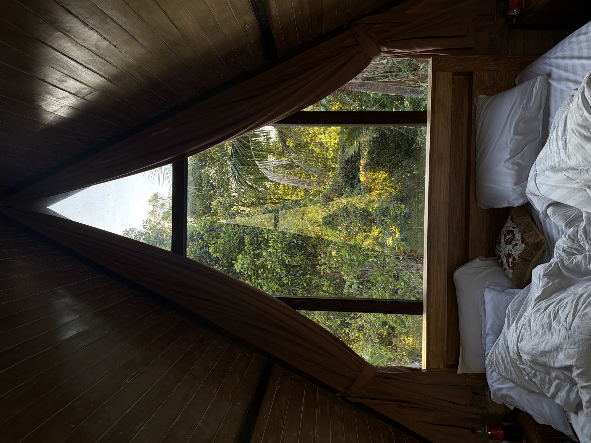 In Sawan, Indonesia, a bedroom with wooden A-frame walls and a bed is depicted. The bed is made with white linens and several pillows. A large triangular window overlooks a lush green forest with sunlight filtering through the leaves. Brown curtains are drawn to the sides of the window.