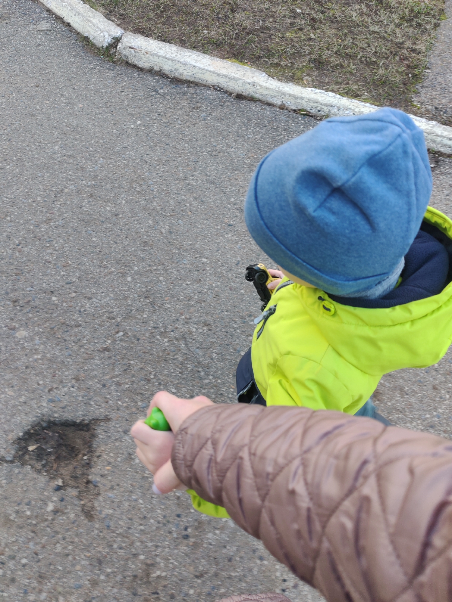 The image, captured from an elevated perspective looking down, features a child and an adult outdoors. A child, whose head and upper body are visible on the right side of the frame, is wearing a blue knit hat and a bright yellow-green jacket over a dark blue inner layer. They are holding a small black and yellow toy car in their hand. An adult's arm, clad in a brown quilted jacket, is visible in the foreground on the left, holding a bright green object, possibly a toy or a small spray bottle.
The setting is an outdoor paved area, likely an asphalt path or sidewalk, with a concrete curb running along its edge. Beyond the curb, there is a patch of dry, sparse grass and dirt. A small, dark, irregularly shaped mark, possibly a wet spot or stain, is visible on the asphalt.
The activity depicts an adult and child walking or being guided. The child is engaged with their toy car. The clothing suggests cool or mild weather, typical for an overcast day, which aligns with the soft, diffused lighting present. No text is visible in the image. The location is in Chaikovsky, Russia.