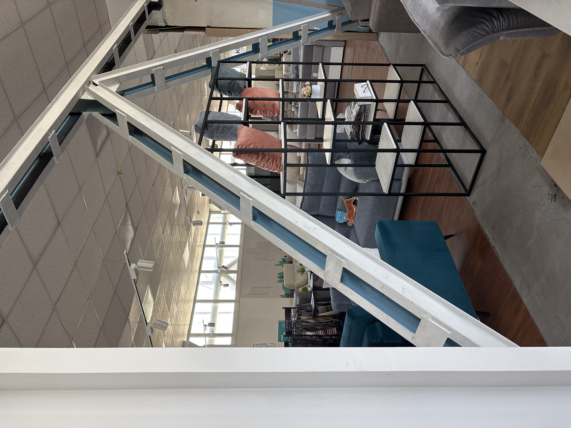 The image depicts the interior of a furniture store. Prominently featured is a large, white metal truss structure with blue accents, extending from the foreground towards the ceiling. Below the truss, to the left, is a teal upholstered sofa. In the mid-ground, a black metal shelving unit displays several cushions, including orange and grey ones, and a small floral arrangement. Behind the shelving unit, several sofas and seating arrangements are visible, suggesting a showroom environment. The ceiling is a dropped tile ceiling with recessed lighting. Large windows line the back wall, allowing natural light to enter. The floor appears to be a mix of concrete and wooden planks. A rack of fabric samples is visible on the far left, and various furniture pieces are scattered throughout the space. There are no people visible in the image. The location is identified as a furniture store in Zaporizhzhia, Ukraine.