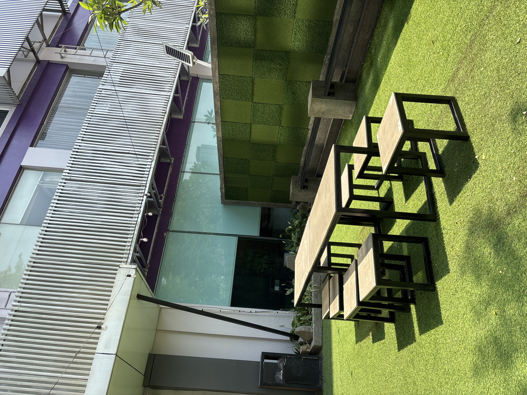 A high-angle, medium shot captures an outdoor seating area on artificial turf at a modern building in Binong, Indonesia. In the foreground, a long, rectangular wooden table with a light brown top and black metal legs is surrounded by six small, square stools with matching black legs and light brown tops. The stools are arranged in pairs on each side of the table. The artificial turf is a vibrant green, with distinct shadows cast by the table and stools due to the bright sunlight.
Behind the seating area, a wall covered in a grid of dark and light green textured panels creates a decorative backdrop. Two concrete blocks with metal railings are positioned in front of this wall. To the left of the table, a black metal trash receptacle with a grey top is situated next to some rocks and low-lying plants.
The background reveals a multi-story building with a facade featuring a series of vertical silver fins. Large windows with light blue blinds are visible on the upper floors, and a purple horizontal band runs across the building above the windows. String lights with exposed bulbs hang from wires strung across the outdoor space. The environment suggests a commercial or hospitality setting, possibly a cafe or restaurant with an outdoor seating option. The overall scene is clean and well-maintained.
