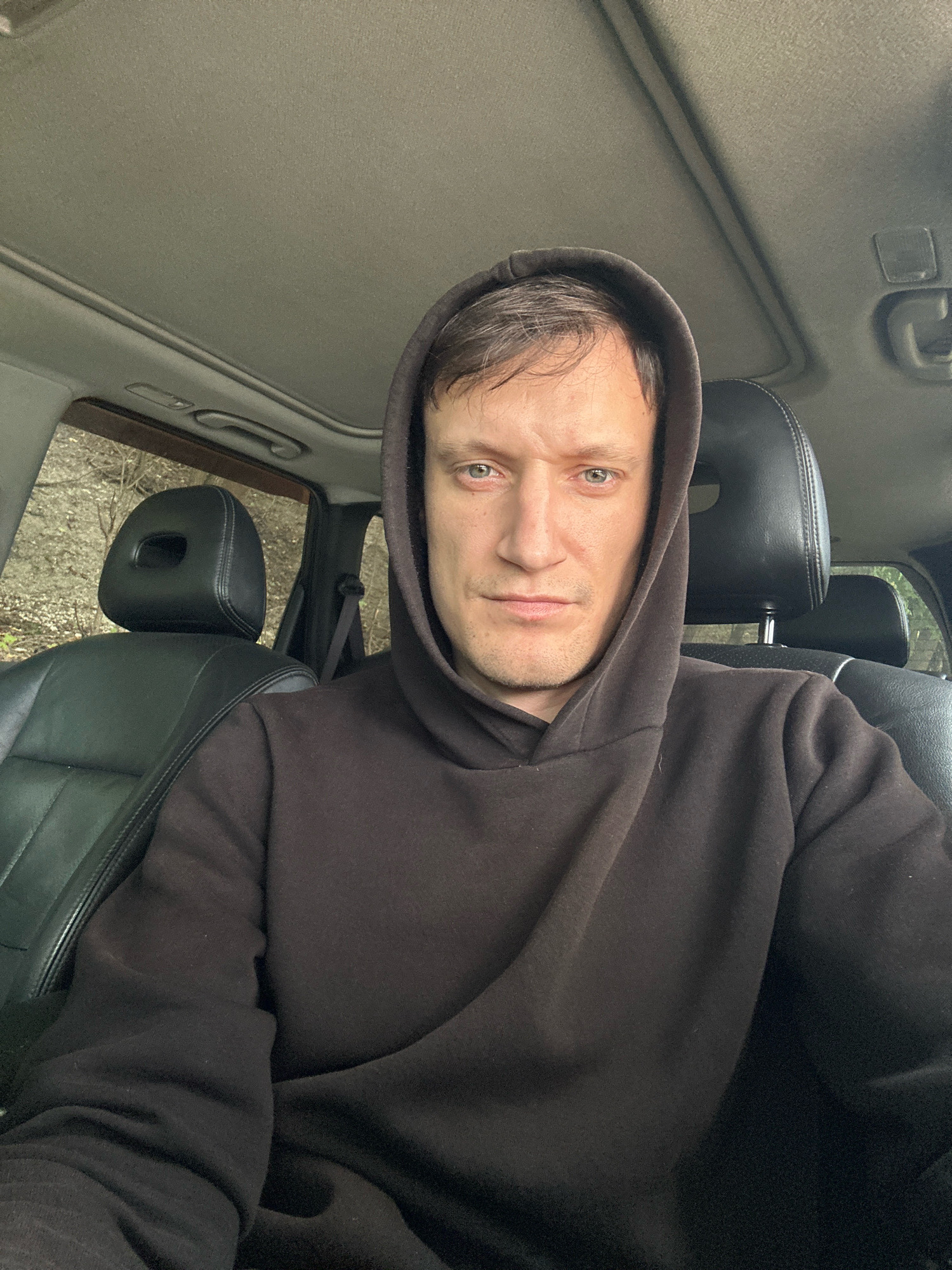 A man wearing a dark brown hooded sweatshirt sits in the driver's seat of a car. He has light-colored eyes and short, brown hair. The car's interior is visible, including the black leather seats, the sunroof, and the dashboard. Outside the car, trees and foliage can be seen through the windows. The scene appears to be set in a natural environment, possibly a rural area or a park. The man is looking directly at the camera, and his expression is neutral. There are no other people or objects of note in the image. The location is not specified as Makiv, Ukraine.