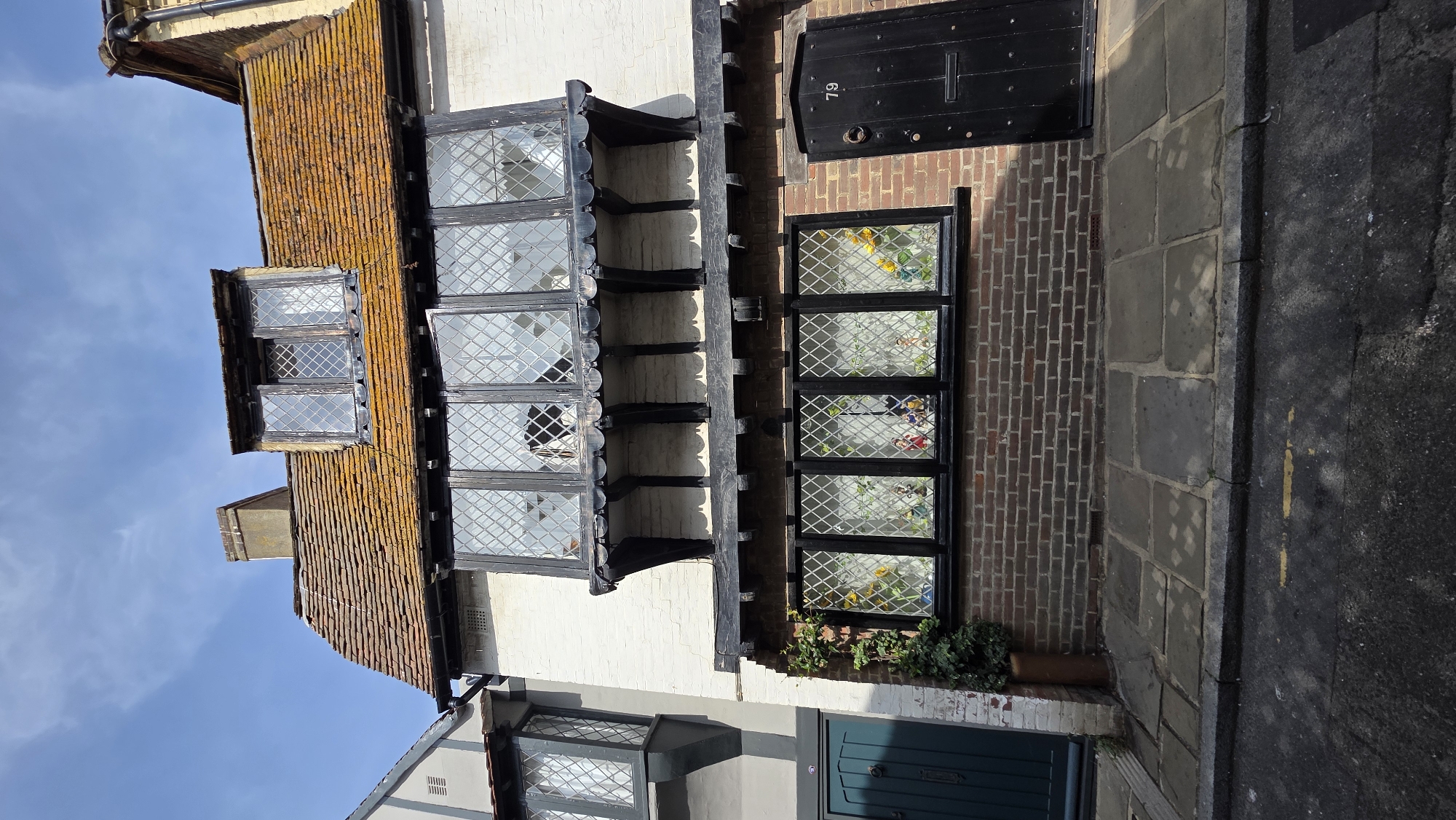 This is a full-frame, eye-level, medium-shot vertical photograph of the exterior of a building at 79 Norman Road, St Leonards, United Kingdom. The building is a narrow, two-story structure with a pitched, orange-tiled roof that has moss and lichen growth. The upper facade is white-washed brick with black wooden trim. There is a large, projecting bay window on the second story, composed of multiple diamond-paned windows. Above this, a dormer window with similar diamond panes is set into the roof. The lower facade is brick with a black wooden door on the right, featuring a round knocker and the number "79" above it. To the left of the door is a smaller, ground-floor window, also with diamond panes, displaying colorful figures and objects inside. A dark green door is visible on the adjacent building to the left, which has a lighter-colored facade and a projecting bay window. The ground in front of the building is paved with large flagstones, bordered by a darker stone curb, and a paved road runs in front of that. Sunlight casts shadows from the building onto the pavement. The sky above is blue with scattered white clouds.