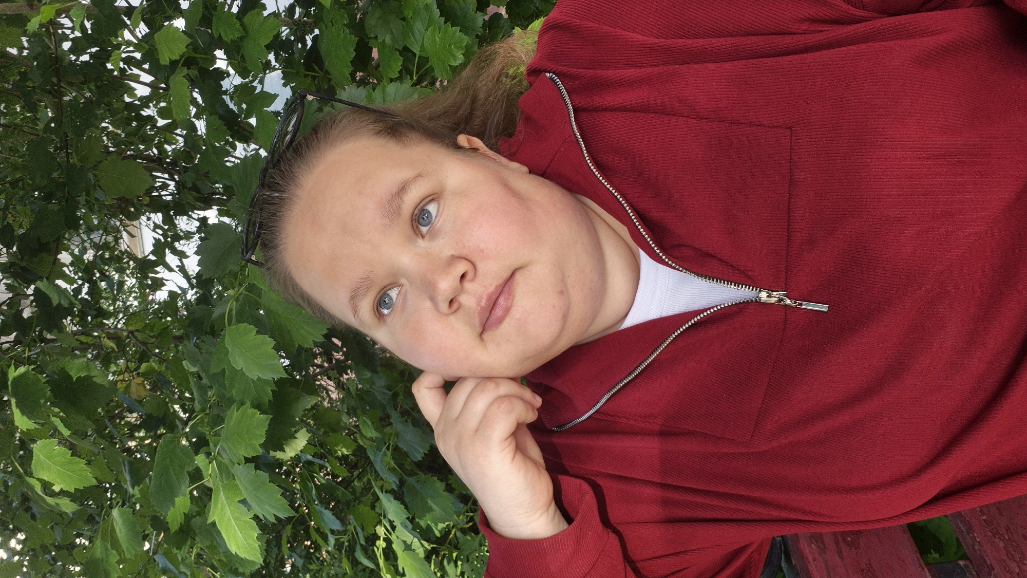 A fair-skinned woman with blue eyes and light brown hair is posing for a selfie outdoors. She is wearing a red, half-zipped sweatshirt over a white top. Her left hand is gently touching her cheek. A pair of glasses are perched on top of her head. Lush green foliage is behind and to the left of her. A wooden bench is visible in the bottom right corner of the image. The photo was taken in the city of Chaikovsky, Russia.