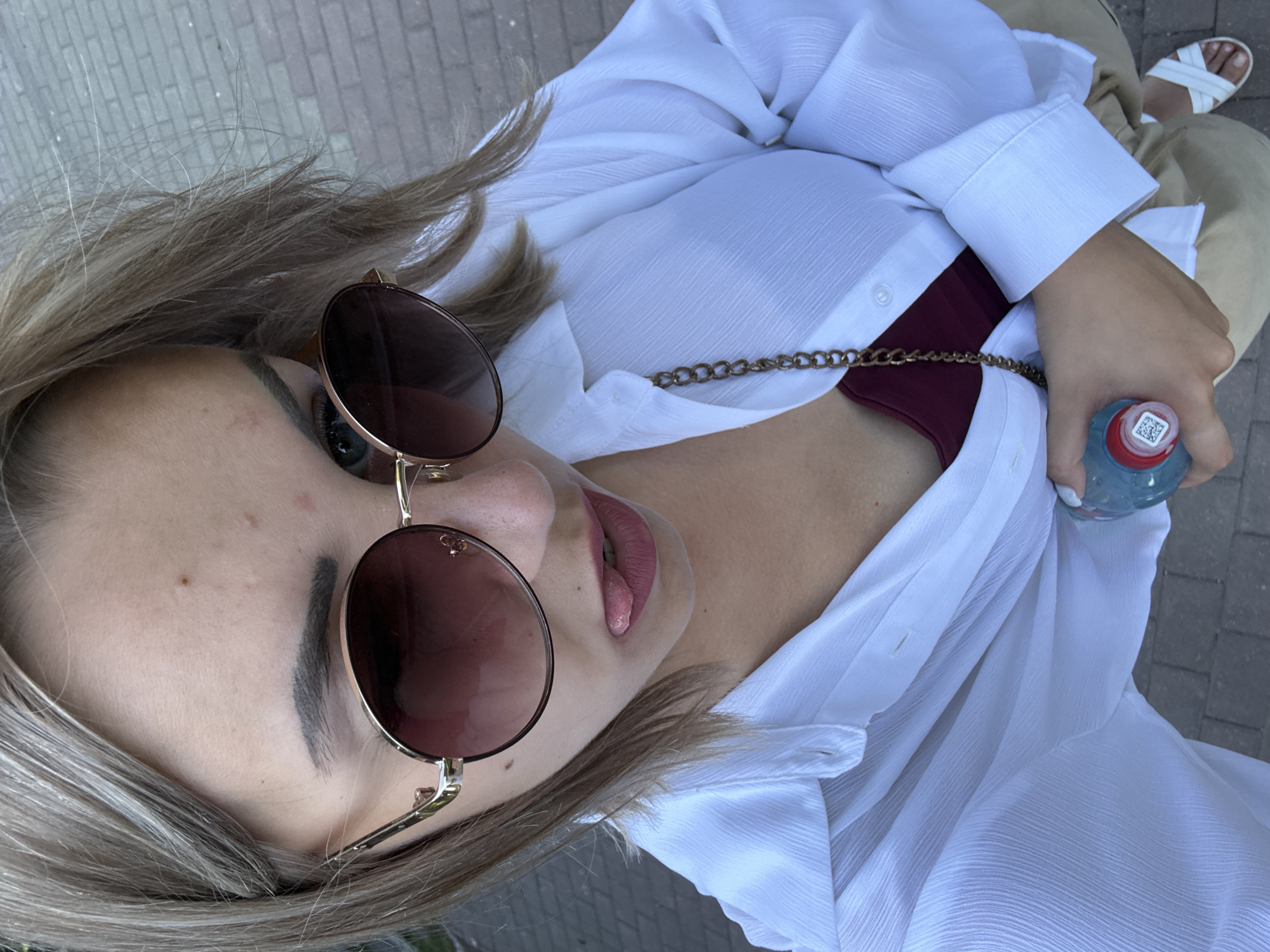 The image shows a close-up, high-angle shot of a young woman outdoors, possibly in Yuzhno-Sakhalinsk, Russia, given the context of the request. She has light-colored hair and is wearing oversized, round sunglasses with a gradient lens. Her lips are painted with a matte lipstick, and her tongue is sticking out. She is wearing a white, unbuttoned collared shirt over a dark red tank top. A gold chain purse strap is visible across her chest. In her right hand, she holds a clear plastic bottle with a blue liquid and a white label. She is also wearing tan pants and white sandals. The background appears to be a paved walkway made of brick-like tiles.