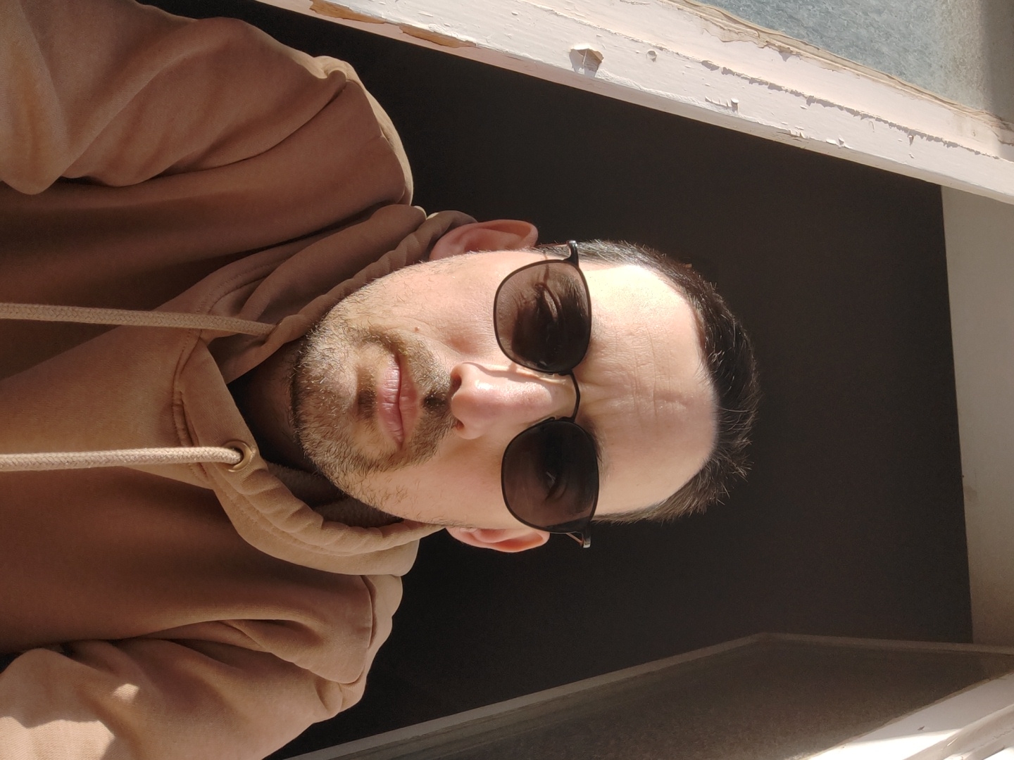 A man wearing sunglasses and a light brown hoodie stands in a window frame. He has short dark hair, a beard, and is looking at the camera. The window frame is painted white and has some chipped paint. Behind him is a dark background, likely the interior of a room. The location is not identifiable as Tel-Aviv, Israel based on the provided image.