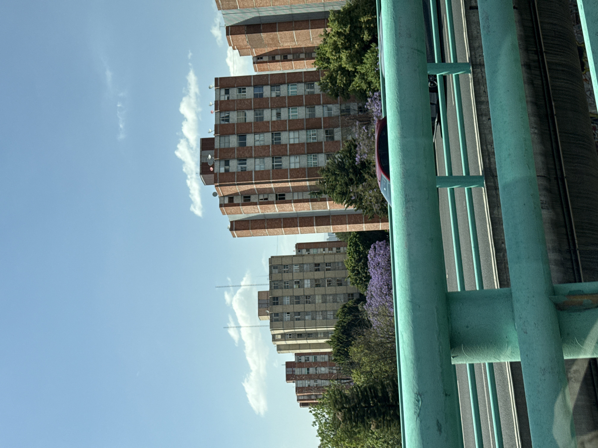 The image shows a view from a bridge in Mexico City, Mexico. In the background are several high-rise buildings with repetitive window patterns, suggesting residential or office use. The buildings have a mix of light gray and reddish-brown facades. Some trees and flowering bushes partially obscure the buildings. A clear, blue sky with some wispy clouds is visible above the buildings. In the foreground, there's a green painted metal bridge structure with vertical and horizontal elements. A highway runs below, and a car is visible on the road. A concrete support structure is partially visible alongside the highway.