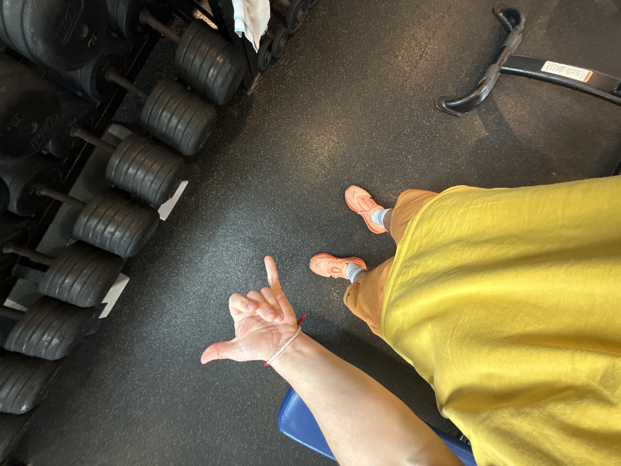 A view from above depicts a person in a gym. The person is wearing orange running shoes with white socks, tan shorts, and a yellow shirt. They are seated on a blue bench. Their left hand is raised, making the "hang loose" gesture. To the left, there's a rack of black weights. A piece of gym equipment is visible in the upper right corner. The gym floor is dark gray. The location cannot be determined to be in Provincetown, United States of America without additional information.