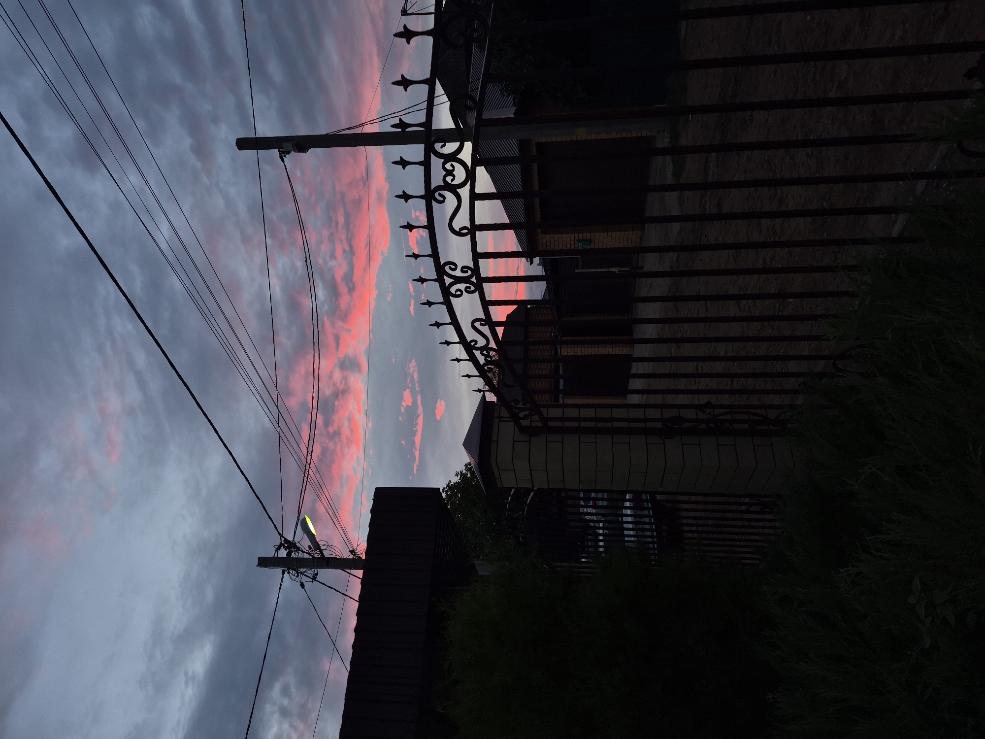 The image shows a street with houses and a fence at dusk. The sky is filled with clouds, some of which are lit with pink and orange hues from the setting sun. There are power lines and a street light visible. The houses appear to be single-family dwellings with garages. The fence is made of metal and has a decorative top. In the foreground, there is a dark, shadowy bush. The overall impression is that of a residential area at the end of the day. The specific location in Astrakhan, Russia, cannot be determined from the image alone, as there are no identifying landmarks or features.