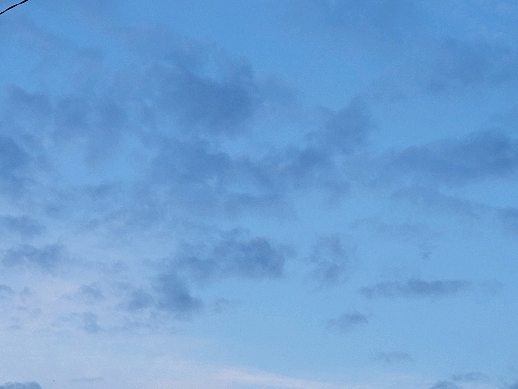 The image displays a wide expanse of a pale blue sky with scattered, wispy grey clouds. The lighting suggests it might be early morning or late evening, with the sun not yet fully risen or already set. No identifiable landmarks of Mežciems, Latvia, such as buildings, streets, or trees, are visible, as the frame is entirely occupied by the sky. There are no people, subjects, objects, or actions depicted in the scene. The overall environment is serene and atmospheric, characterized solely by the celestial view.