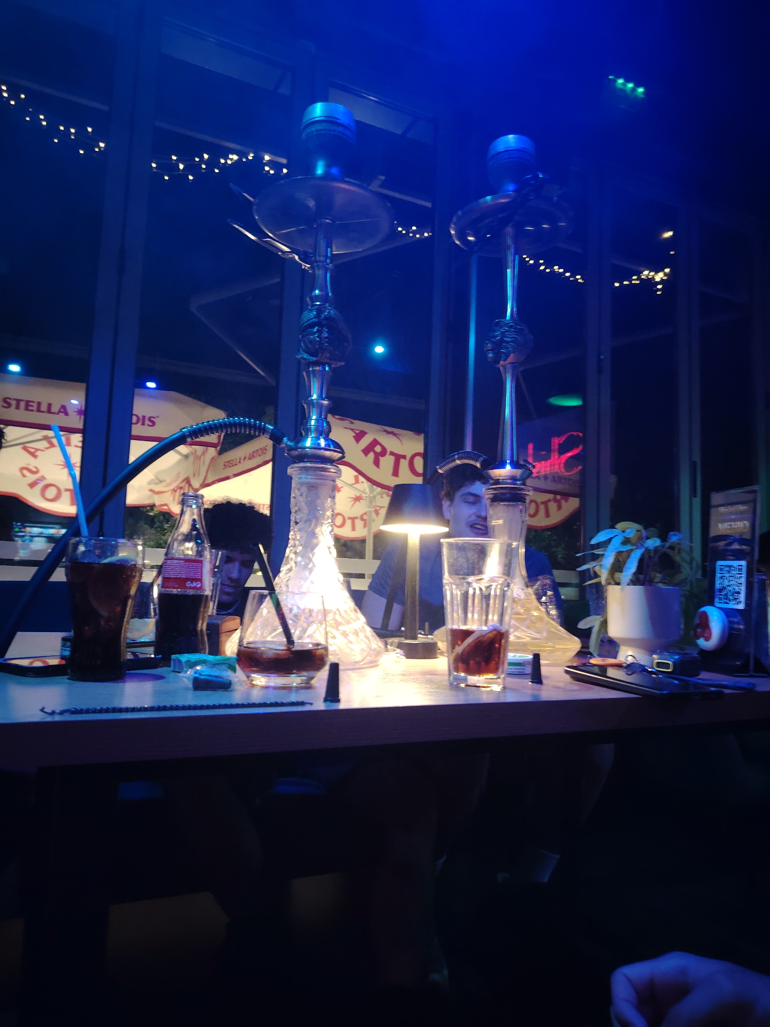 This image captures a dimly lit, possibly indoor, setting with blue lighting. The foreground features a wooden table laden with several items. To the left, a tall glass filled with a dark beverage and a blue straw stands next to a bottle of Coca-Cola. Further right, another glass contains a darker liquid with ice and a straw. A shisha pipe, a common fixture in hookah lounges, is prominently displayed in the middle of the table, with its ornate metal stem and glass base. Behind the table, two individuals are partially visible. One person, with dark, curly hair, appears to be looking down, while another person sits behind them, obscured by the shisha pipe. In the background, through a large window or glass partition, a string of lights illuminates the exterior. In the distance, large white umbrellas with red lettering, possibly advertising "STELLA" and "ARTOS," are visible, suggesting an outdoor seating area or a view of another establishment. The overall ambiance suggests a relaxed, social environment, likely a bar or lounge. The presence of shisha pipes and the advertising banners may indicate the location is a specific type of establishment. Without more context, it's difficult to pinpoint the exact location, but the mention of Plovdiv, Bulgaria, along with the visual cues of outdoor seating and prominent brand advertising, suggests a venue that caters to patrons enjoying drinks and shisha, possibly in an urban setting.
