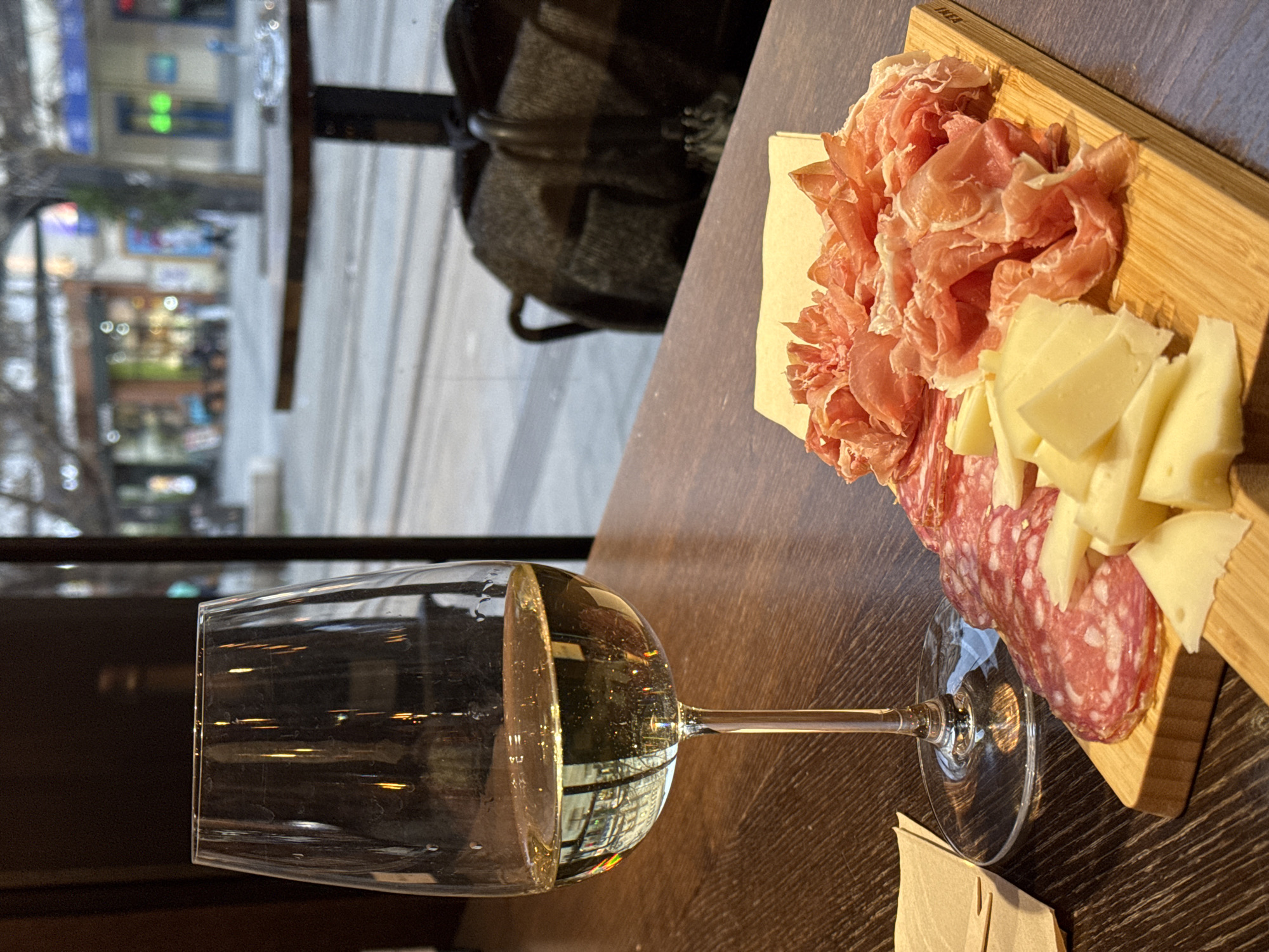 The image shows a table setting in what appears to be a restaurant or bar, likely in Sofia, Bulgaria. A wine glass, filled with a light-colored liquid, possibly white wine, is placed in the foreground. Next to the glass is a wooden board with an assortment of cured meats and cheese. There is prosciutto-like meat, sliced salami, and cubed white cheese arranged on the board. There is a paper napkin next to the cheese board. The table is dark wood. In the background, a window provides a blurred view of a street scene. Buildings and lights are visible across the street. A chair is visible between the table and the window. The scene suggests a social setting.