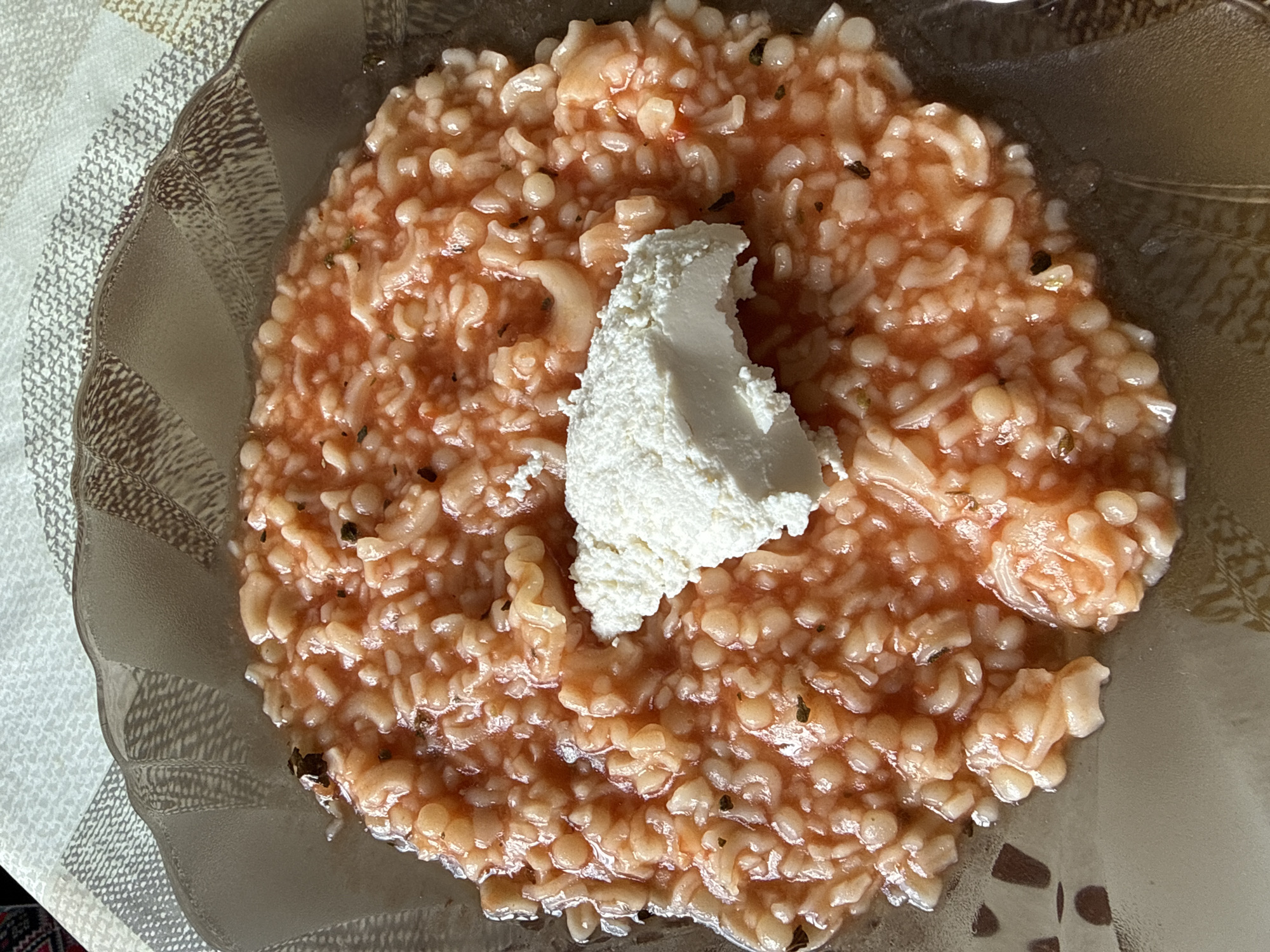 A top-down close-up shot shows a clear glass bowl filled with orzo pasta in a reddish sauce. A dollop of white cheese sits in the center of the pasta. The bowl is placed on a patterned placemat.
