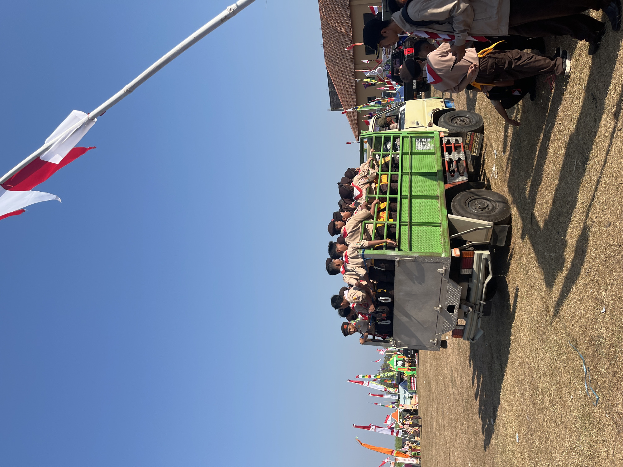 A group of scouts, dressed in uniforms consisting of brown shirts and pants, are riding in the back of a light-colored truck with a green metal bed. The truck is parked on a dry, grassy field under a clear blue sky. Several flags are visible in the background, including red and white flags, suggesting a festive or official event. Some scouts are looking out, while others appear to be talking amongst themselves. The scene appears to be taking place outdoors, possibly at a rally or campground. The truck has license plate number "W 9548 A". A building with an orange-tiled roof is visible in the far background. This event likely took place in the city of Gembong, Indonesia.