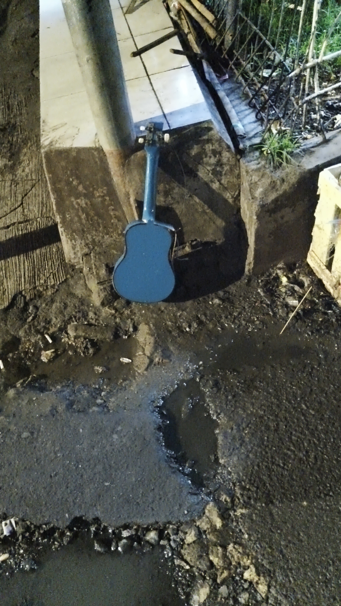 An overhead shot depicts a blue ukulele leaning against a concrete pillar in South Jakarta, Indonesia. The ukulele is positioned on a patch of broken asphalt and dirt, which also contains several cigarette butts and puddles of water. To the right of the ukulele, a metal fence encloses an area with green grass and debris. The scene is dimly lit, suggesting it is nighttime.