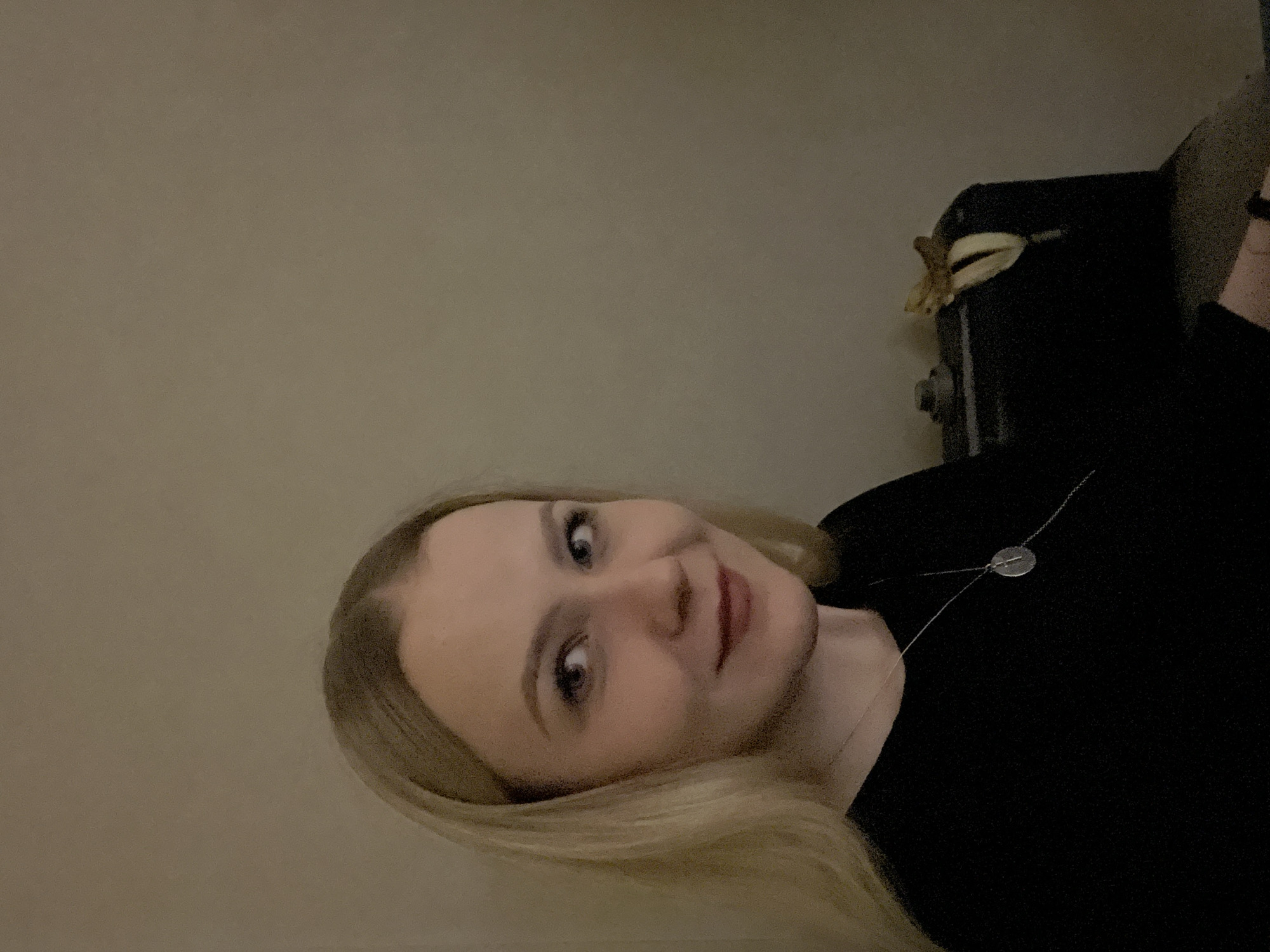 A young woman is captured in a waist-up portrait, looking directly at the camera with a subtle, friendly smile. She has long, blonde hair, fair skin, and light-colored eyes. She wears dark, likely black, clothing and a delicate silver-toned necklace with a small, circular pendant.
The background is simple, featuring a plain, light brown or beige wall on the left side. On the right, behind her right shoulder, a dark object—possibly a piece of furniture or a box—is visible. On top of this dark object, there is a light-colored, abstract item that resembles a dried banana peel or a decorative bow.
The lighting is dim, suggesting an indoor setting with low or artificial light, possibly in the evening or at night. The image has a slightly grainy texture. No visible text is present. The overall atmosphere is calm and casual. The location is in Khimki, Russia.