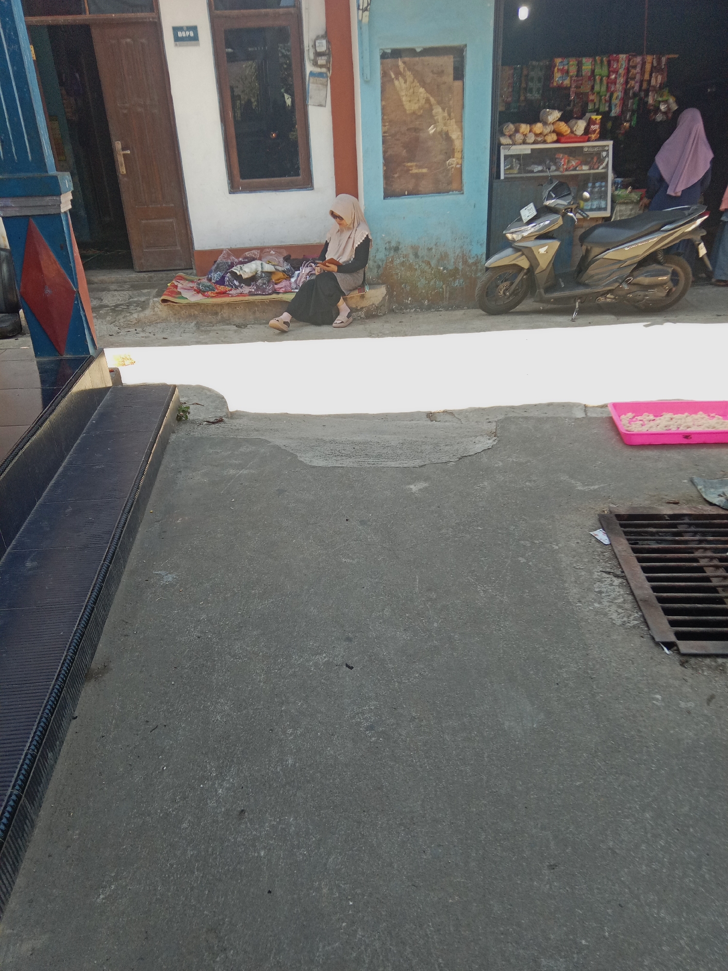 The image depicts a scene in front of a small store in Watukumpul, Indonesia. A young woman wearing a beige hijab and a dark long-sleeved dress is sitting on the ground, facing a pile of colorful fabrics. She is holding a dark object, possibly a mobile phone, in her hands and appears to be engaged with it. Beside her, a motorcycle is parked on the street. The store behind her displays various goods on shelves and hanging racks. Another woman, wearing a pink hijab, is visible inside the store. The ground in front of the store is paved with concrete, and there is a metal grate on the right side of the image. A pink tray containing small white objects is also placed on the concrete. A set of dark tiled stairs is visible on the left side of the image, partially obscuring a blue and red pillar. The overall environment suggests a typical street scene in a local Indonesian community.