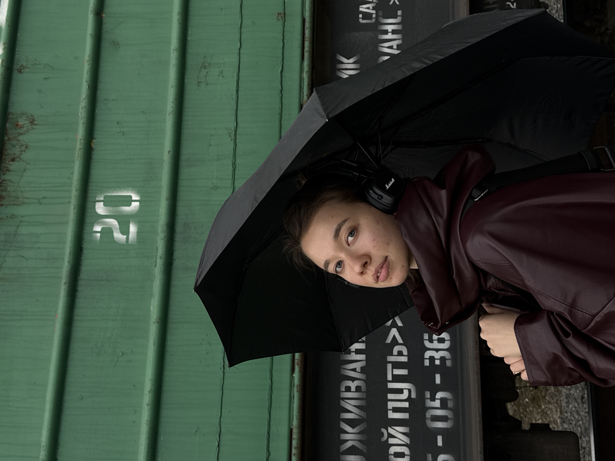 A young woman stands in front of a green and black train car in Chaikovsky, Russia. She is holding a black umbrella and wearing a maroon coat and black headphones. The number "20" is stenciled in white on the green train car. Russian text is visible on the black train car.