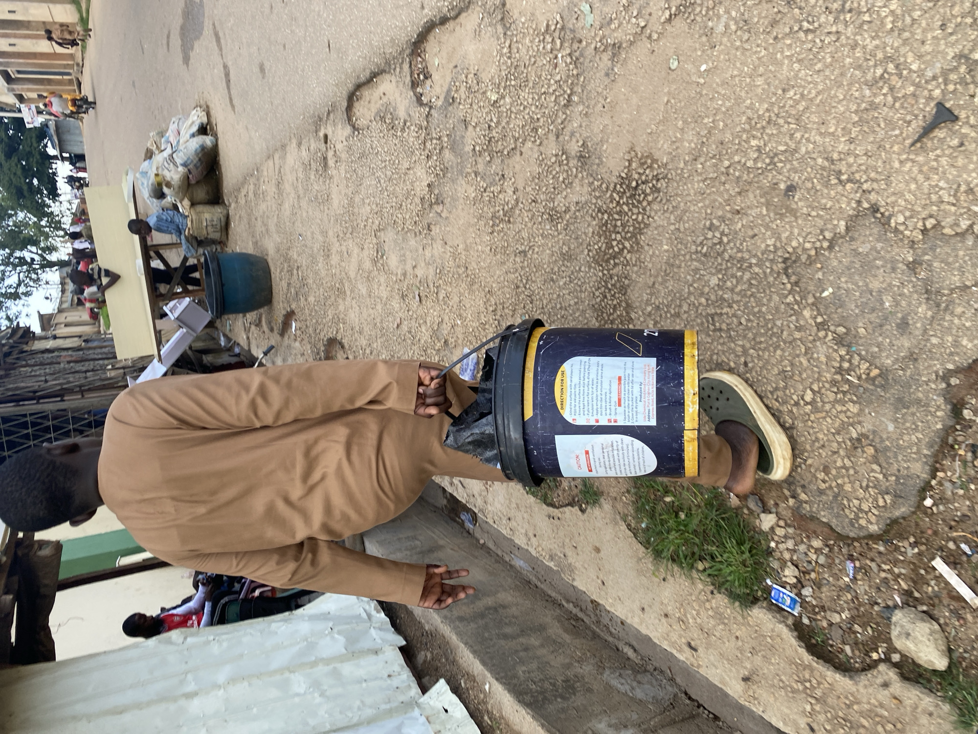 The image depicts a street scene in Jos, Nigeria, with a man carrying a bucket of paint and walking on a cracked asphalt road. The man is wearing a brown tunic and dark sandals. The bucket has a yellow lid and a label with instructions for use. In the background, other individuals are visible, including a person on a motorcycle and others near a market stall selling goods. The environment shows a mix of residential buildings and street vendors, indicative of a bustling urban area. There are also some debris and potholes on the road.