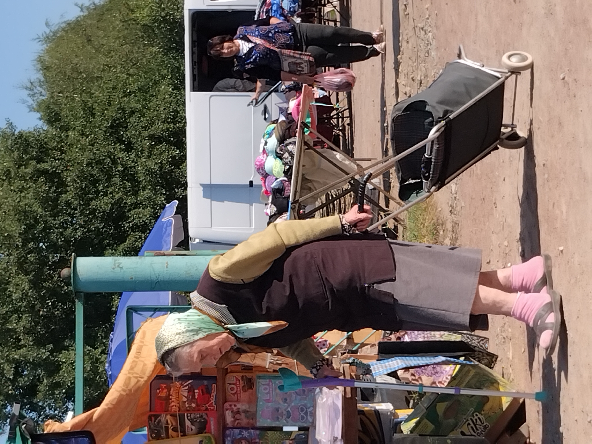 In Irshansk, Ukraine, an elderly woman wearing a headscarf, dark vest, and gray skirt, leans on a blue and purple crutch while walking through an outdoor market. She is accompanied by a wheeled shopping cart. To her right, another woman in a patterned vest and dark pants carries a pink shopping bag. In the background, a white van is parked, and trees form a natural backdrop against a clear sky. Stalls with merchandise, including clothing and toys, are visible throughout the scene.