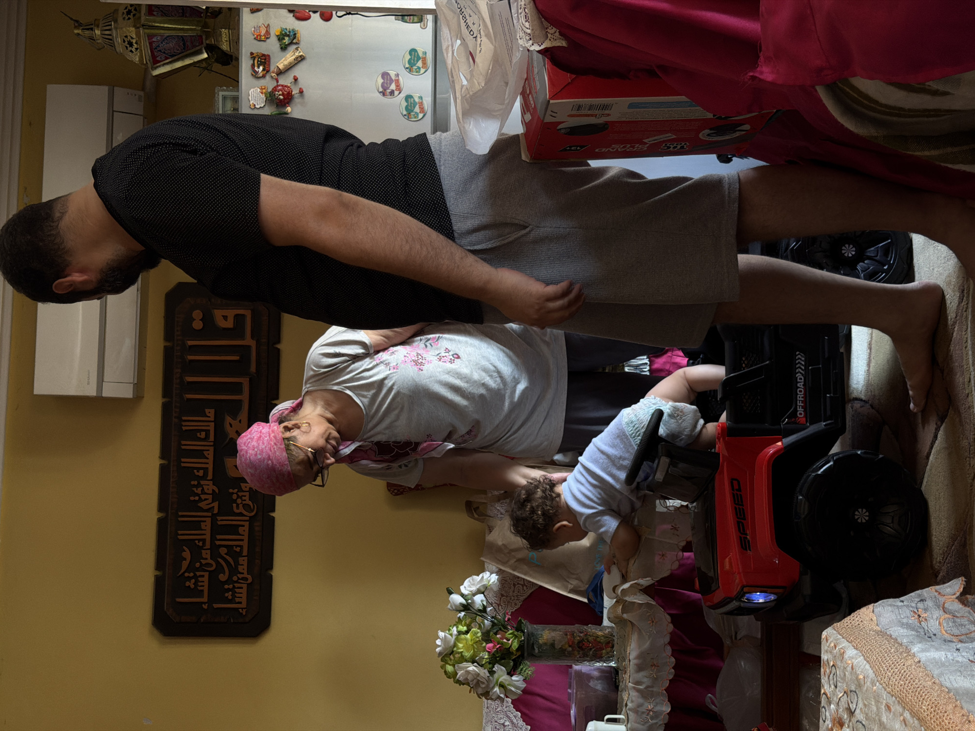 In a room in 6th of October, Egypt, a man and a woman stand over a toddler who is interacting with a red toy Jeep. The man, with a beard, wears a black, dotted t-shirt and grey shorts, standing with his back to the viewer. The woman, wearing glasses and a pink headscarf, has on a grey t-shirt and dark pants. The toddler is on its hands and knees, leaning forward onto the toy Jeep, wearing a diaper. The Jeep has the word "SPEED" printed on its side. The room is decorated with a wooden Arabic calligraphy art piece on the yellow wall, and various items are visible including a vase of flowers on a covered table, a refrigerator with magnets, and a large golden lantern.
