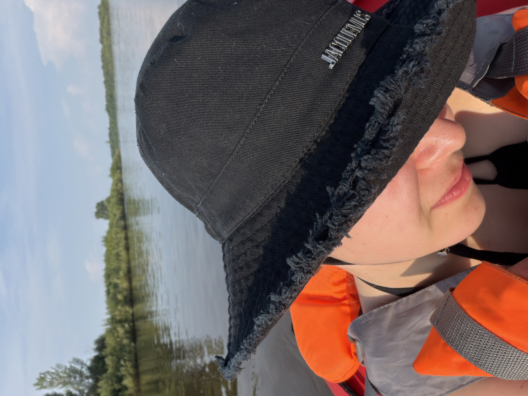 A close-up shot shows a person wearing a black bucket hat and an orange life vest on a body of water. The hat has a frayed edge and a silver "JACQUEMUS" logo on the side. The person's face is partially visible, with their head tilted down. The background shows a calm river with green foliage on the banks and a blue sky with some clouds. The scene appears to be in the city of Kalino, Russia, likely during daylight hours.