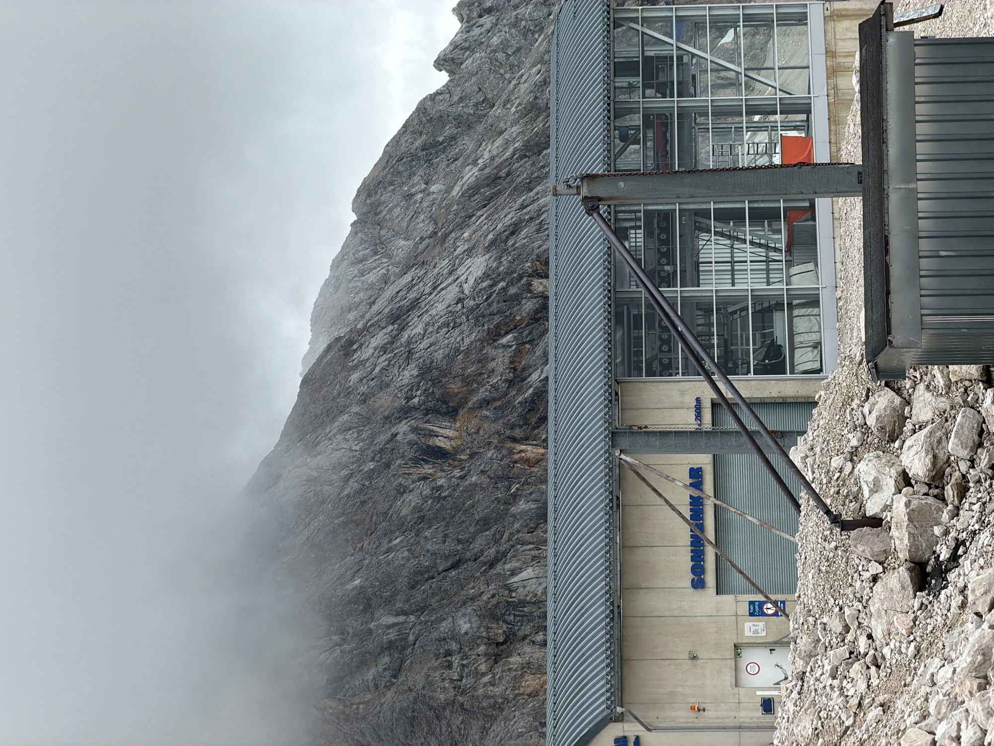 The image depicts a building at a ski resort in Garmisch-Partenkirchen, Germany, likely part of a ski lift station. The building has a grey, corrugated metal roof and a concrete facade. Large windows offer a view of the interior, which appears to house machinery for a ski lift, including cables and gondolas. The word "SONNENKAR" is visible on the facade, along with a sign indicating "2600m". The building is situated on a rocky slope, with large boulders and debris in the foreground. In the background, a large, rugged mountain range is visible, partially obscured by clouds and fog. The sky is overcast, suggesting cool, possibly damp weather. There are no people visible in the image.