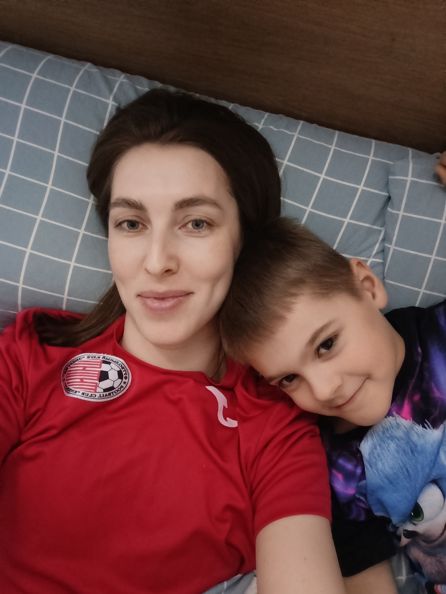 The image captures a close-up selfie of two individuals, an adult female and a young male child, in what appears to be an indoor, domestic setting. The adult female, exhibiting long dark hair, a light complexion, and blue eyes, occupies the left side of the frame, looking directly at the camera with a subtle smile. She is dressed in a red short-sleeved top featuring a circular white patch on the left chest, which contains mirrored Russian text translating to "FOOTBALL CLUB" and a soccer ball graphic. The young male child, estimated to be between 5 and 10 years old with short brown hair and brown eyes, is positioned to the right, leaning his head against the adult female's shoulder/chest area. He is also smiling faintly and looking towards the camera, wearing a dark t-shirt with a partial blue cartoon character graphic, identifiable as Sonic the Hedgehog. Both individuals are lying on a light blue pillow patterned with a white grid, while a dark wooden surface, possibly a headboard, is visible in the upper right background. The specific location within the city of Chaikovsky, Russia, cannot be ascertained from the visual information presented.