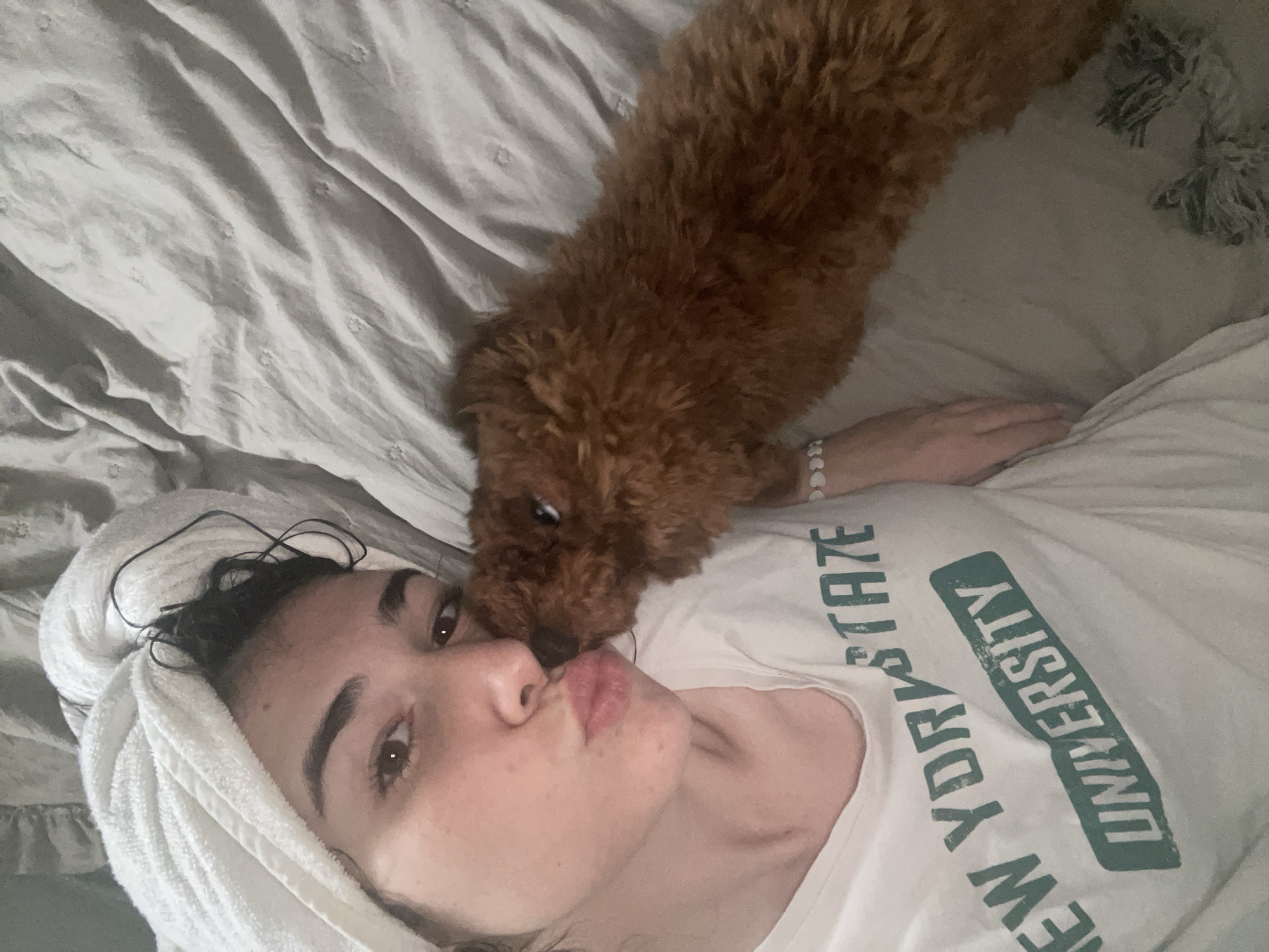 A young woman with dark hair wrapped in a white towel and a small, reddish-brown curly-haired dog are captured in an intimate moment. The woman lies on her back on a light-colored bed with wrinkled sheets, making a kissy face towards the camera. The dog, possibly a poodle or doodle, is positioned above her, leaning down with its snout close to her face, appearing to sniff or offer affection.
The woman wears a white t-shirt with green text, partially visible and reversed, that reads "NEW YORK STATE UNIVERSITY." Her right arm is visible, adorned with a white bracelet featuring heart-shaped beads. Her dark hair beneath the towel appears damp. The dog is entirely reddish-brown with fluffy, curly fur.
The setting appears to be indoors, likely a bedroom, with crumpled light grey or white bedding. In the upper right corner, a portion of a textured, light-colored throw or pillow with tassels is visible. The overall mood of the image is affectionate and relaxed, capturing a tender interaction between the woman and her pet. There are no specific cues for time of day or external weather.