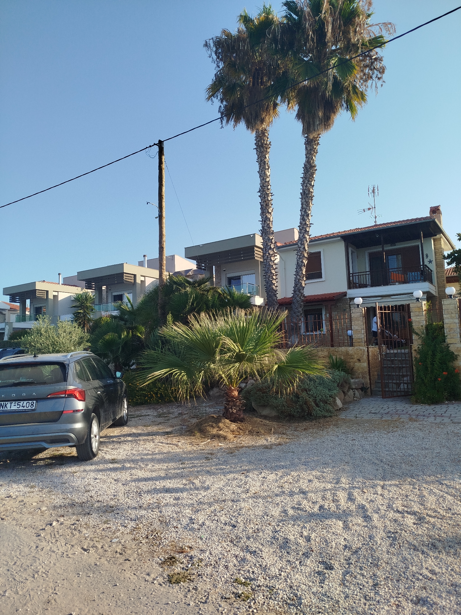 This is a frontal, eye-level, outdoor, full-shot image captured on a sunny day in Sithonia Municipal Unit, Greece. The scene depicts a residential area featuring a row of modern, two-story houses with flat roofs and balconies. The houses are painted in neutral colors, with some featuring darker accents on their upper levels. In front of the houses, there is lush vegetation, including several palm trees of varying sizes and other leafy plants.
A dark gray Skoda station wagon is parked on a gravel driveway in the foreground, facing left. The car's license plate, "NKT 5408," is visible. A wooden utility pole stands to the left of the houses, with power lines running across the frame. The ground is covered in light-colored gravel and sand. A paved pathway leads to an open metal gate in front of the rightmost house, where a person in a white shirt is visible walking. The sky is clear and blue.