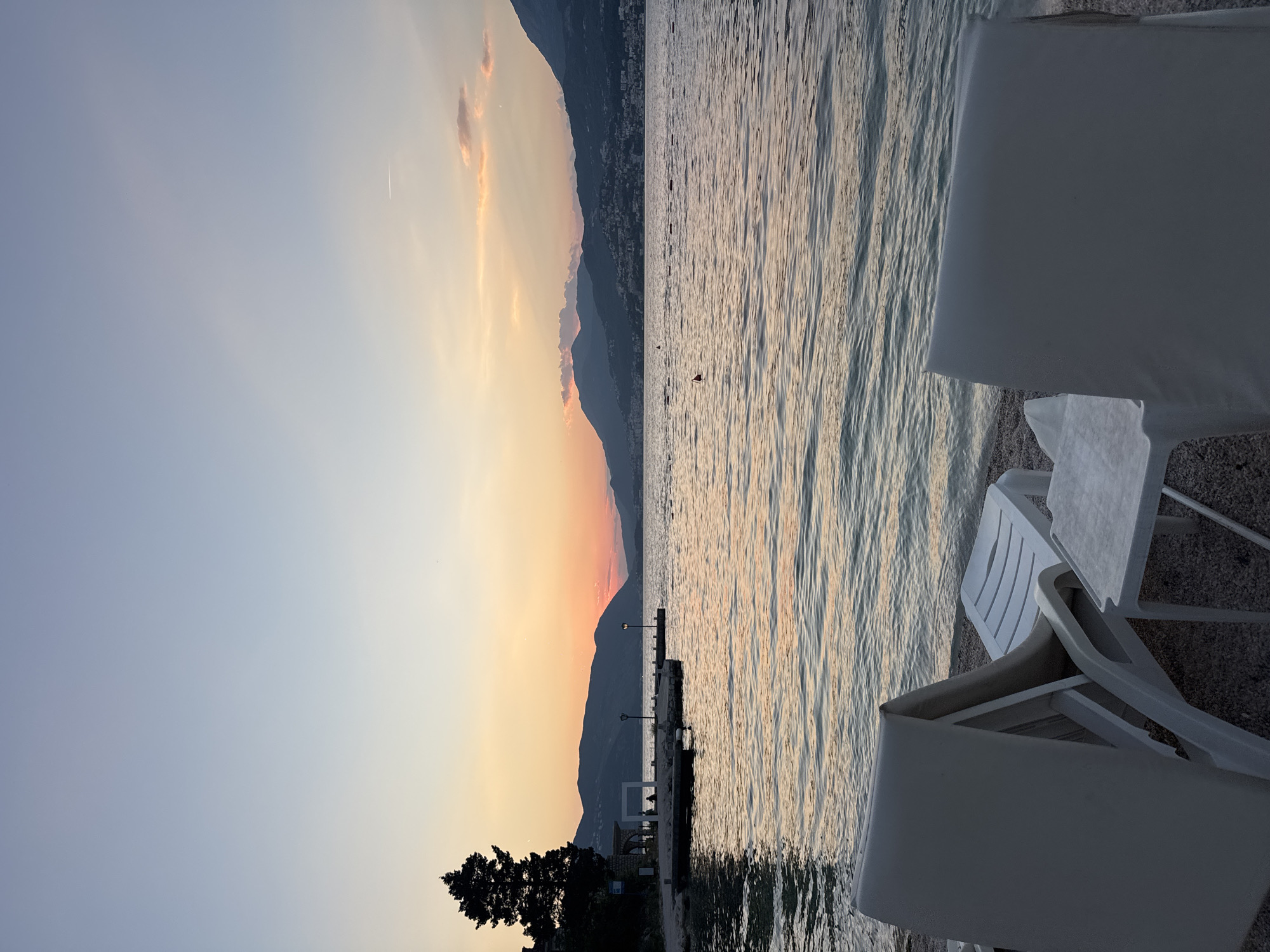 The image depicts a serene beach scene at dusk. Two white lounge chairs and a small white table are positioned on a pebble beach in the foreground. Beyond the beach, a calm body of water reflects the soft hues of the sunset. In the middle ground, a stone pier extends into the water, with a lone tree silhouetted against the sky on the left side of the pier. Further in the distance, mountains form a dark, undulating silhouette against the colorful twilight sky, which is transitioning from pale blue at the top to warm oranges and pinks near the horizon. The overall impression is one of tranquility and the end of a day at the seaside.