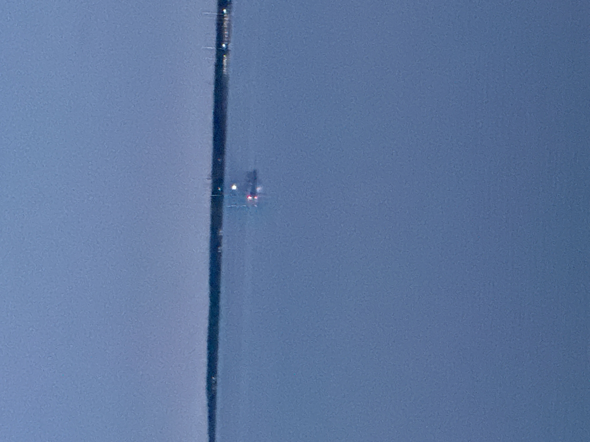 The image shows a view of the sea with a dark line of land in the background. There is a boat in the water, which appears to be illuminated by red and white lights. The sky is a pale blue, suggesting it may be twilight or dawn. The scene is calm and quiet, with no other activity visible. It is not possible to determine the exact location in Tallinn, Estonia from this image alone, but it could be a view from the coast of the city, looking out towards the Baltic Sea.