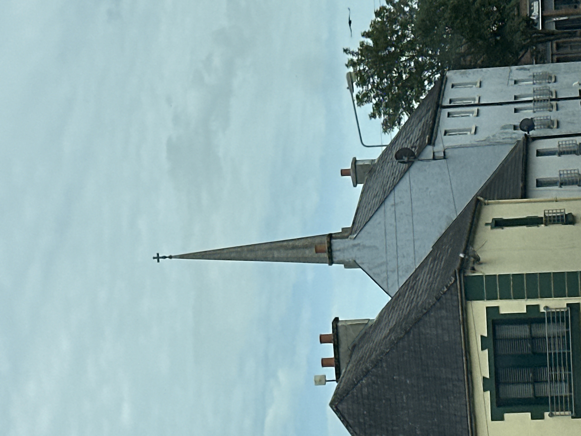 The image is a vertical, outdoor, medium shot of rooftops and a church spire under a cloudy sky. The scene appears to be in a town or city, with the architecture suggesting an urban environment. In the foreground, there's a dark grey roof with multiple chimneys. Behind it, a lighter grey, pointed spire with a cross at its apex extends upwards. The spire is attached to a building with a light grey sloped roof. Further in the background, a multi-story building with numerous windows, some with security bars, is visible. A large green tree with dark foliage is also present, and a lamppost is attached to the side of a building. The sky is overcast and light blue, indicating daytime. There is no visible text in the image. The overall impression is one of a slightly dreary, possibly historic urban landscape. The image's orientation is rotated 90 degrees counter-clockwise.