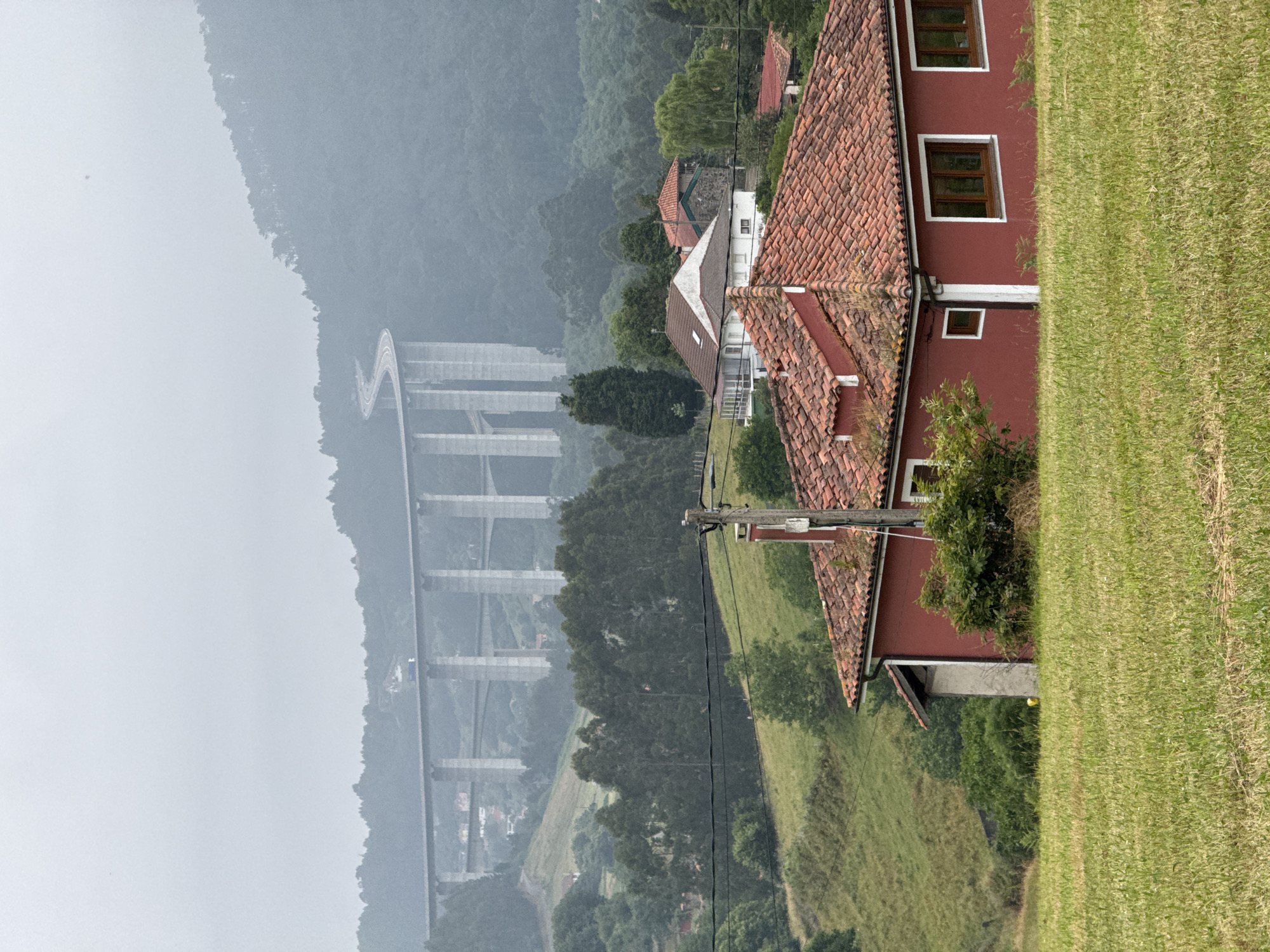 The image captures a rural landscape with a prominent red-tiled roof house in the foreground. Behind it, a winding highway bridge extends across a lush green valley and into the misty, tree-covered hills. The sky is overcast and hazy, lending a muted tone to the scene. The house features classic terracotta roof tiles and red-painted walls with white trim around the windows and a small square window. A power pole with some wiring is visible near the house. In the distance, other houses and trees dot the undulating terrain, and the large concrete structure of the highway bridge dominates the upper portion of the image. The scene appears to be a natural setting with man-made infrastructure integrated into the landscape, likely in the vicinity of Cudillero, Spain, known for its dramatic coastal geography and mountainous terrain.