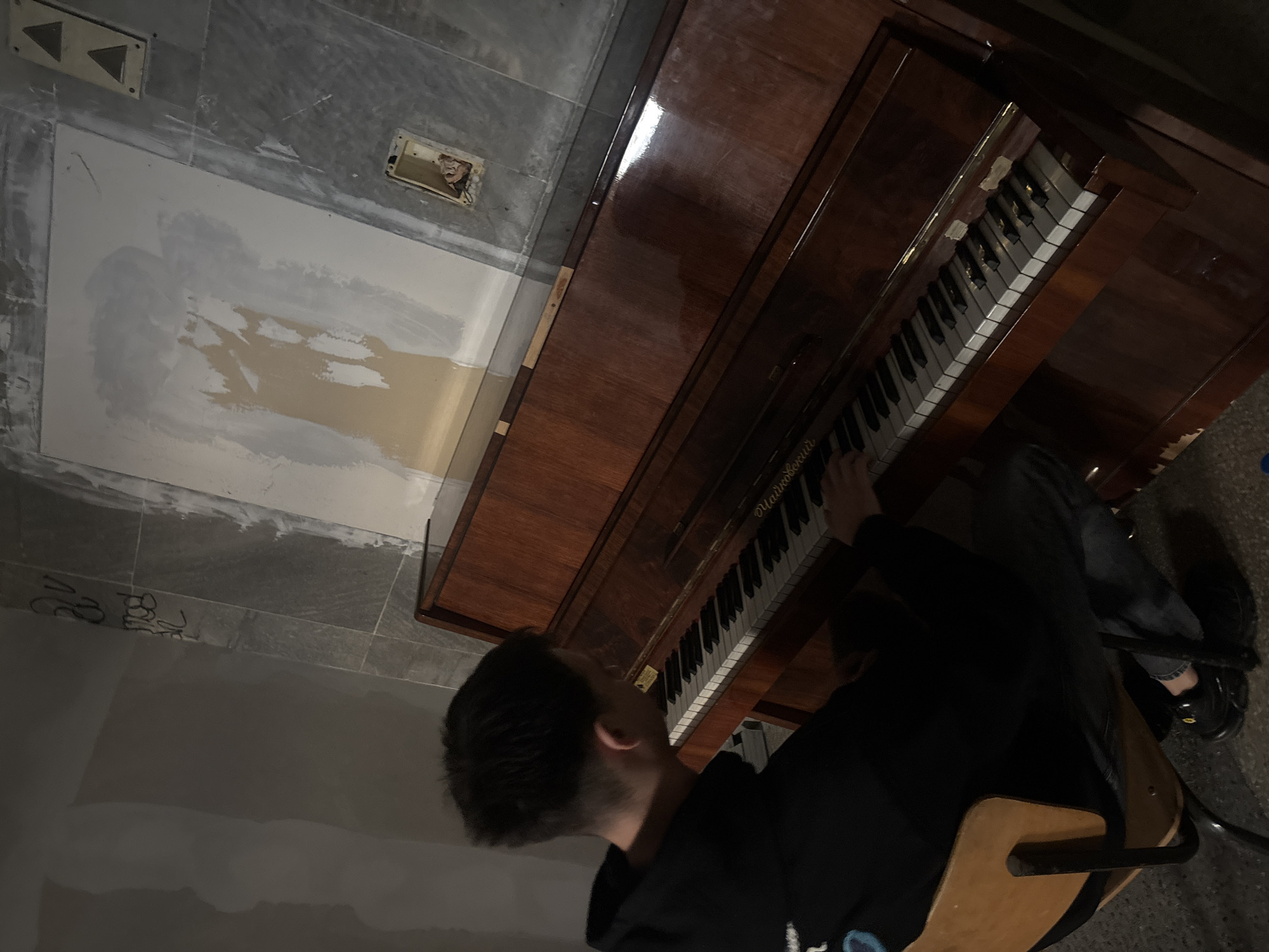 An overhead shot captures a person playing an upright piano in an interior space that appears to be under renovation or minimally furnished.
The main subject is a person, likely male, with dark hair, wearing a dark hoodie and dark jeans. He is seated on a simple wooden chair with a dark metal frame, positioned in front of the piano with his hands on the keys, indicating he is playing.
The piano is a dark brown, glossy wooden upright model. The keyboard features both white and black keys, some of which exhibit signs of wear or discoloration. The name "Чайковский" (Tchaikovsky) is visible in Cyrillic script on the piano's fallboard.
The surrounding environment features a floor with large grey tiles in some areas, while other parts are covered with light-colored plaster or paint, suggesting an unfinished or utilitarian room. A section of the wall behind the piano is also patched with light grey and beige plaster. Two electrical fittings are visible on the wall; one has two triangular buttons, and another is an exposed, damaged outlet with visible wiring. Faint, dark markings, possibly numbers like "32" and "18," are scrawled on the tiled floor.
The lighting in the room is dim, suggesting artificial light. While the image itself doesn't provide explicit location cues, the provided context places the scene in Plovdiv, Bulgaria. The person's face is not visible, so no specific emotions can be discerned.